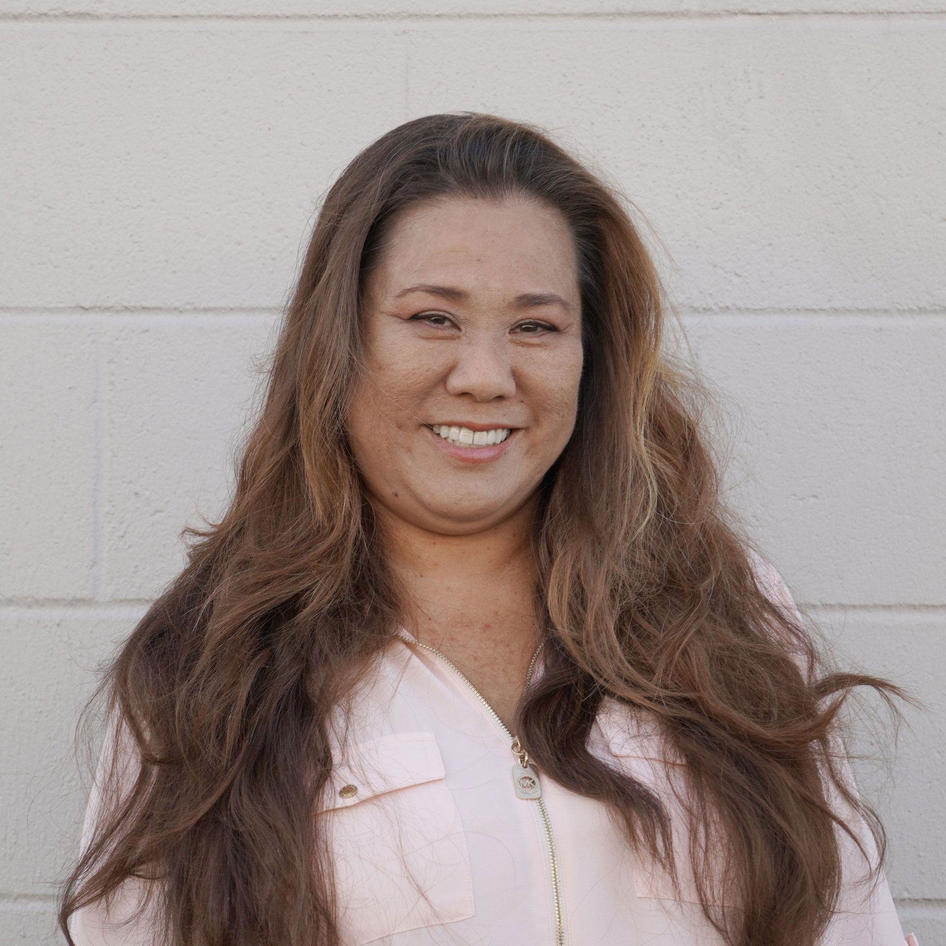 Woman smiling, long brown hair, wearing a pink top, against a white brick wall.