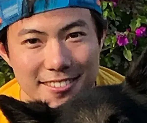 Man smiling, wearing a blue hat, yellow jacket, with a black dog partly visible.