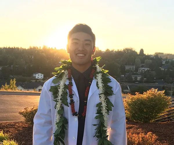 Man in white coat and lei smiles at sunset, overlooking a town.