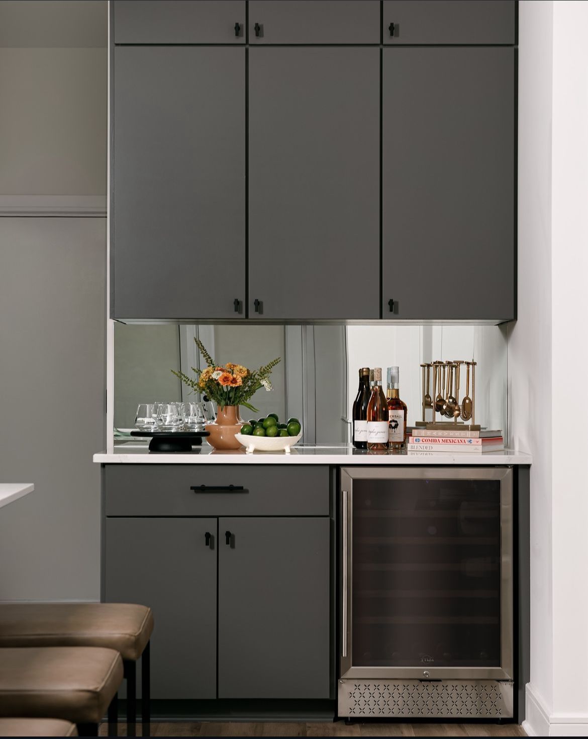 Gray built-in bar cabinet with bottles, glassware, and a small flower arrangement beneath upper storage cabinets.