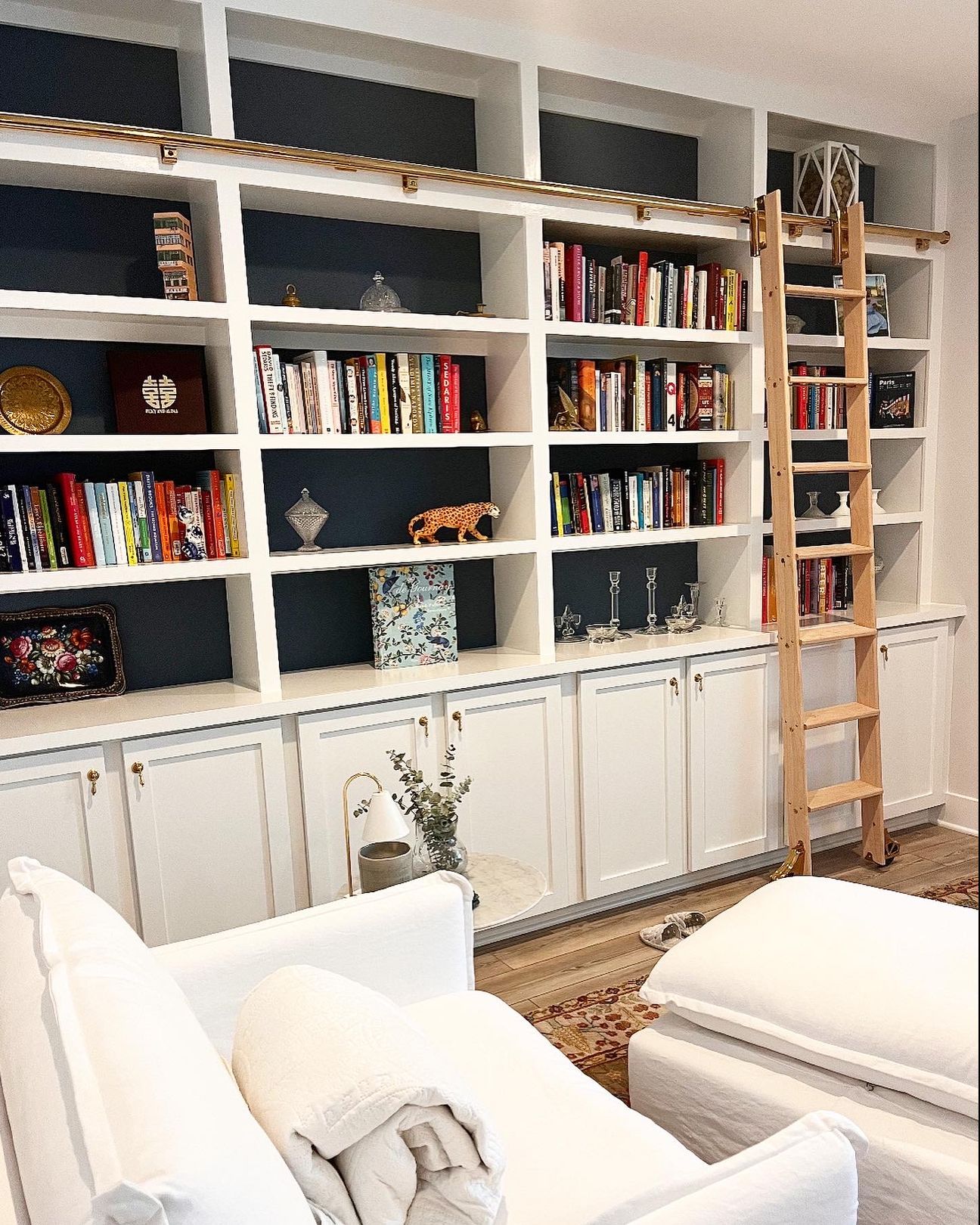 White built-in bookshelves filled with books, a leaning ladder, and white chairs in a bright living room.