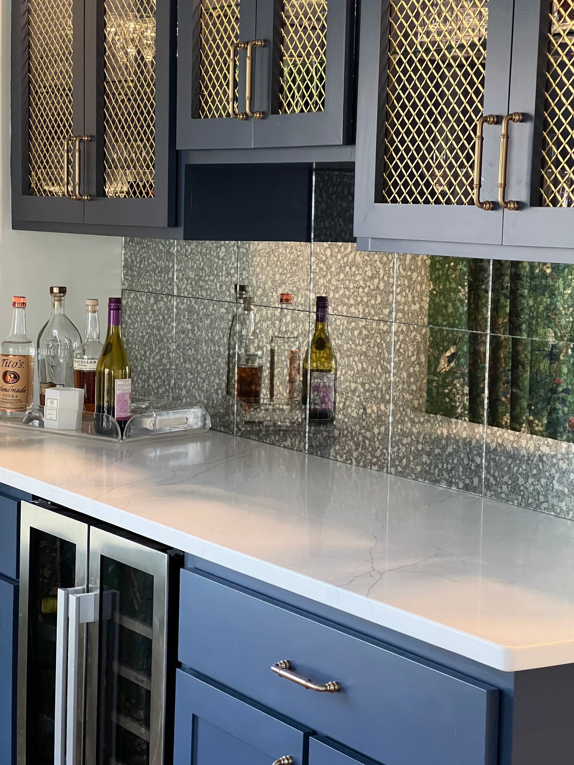 Modern kitchen with blue cabinets, white countertop, mirrored backsplash, and wine bottles displayed above.