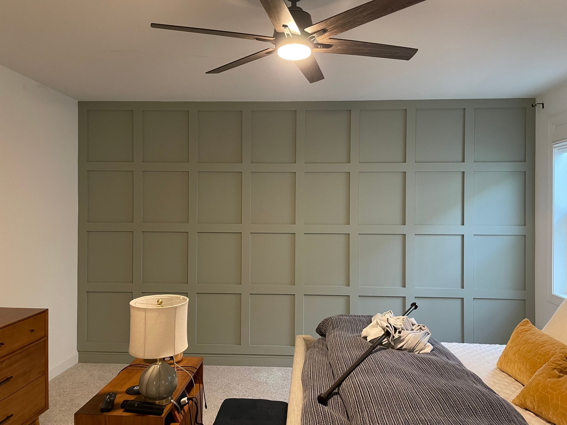 Gray bedroom with paneled accent wall, ceiling fan, bed, dresser, and window light