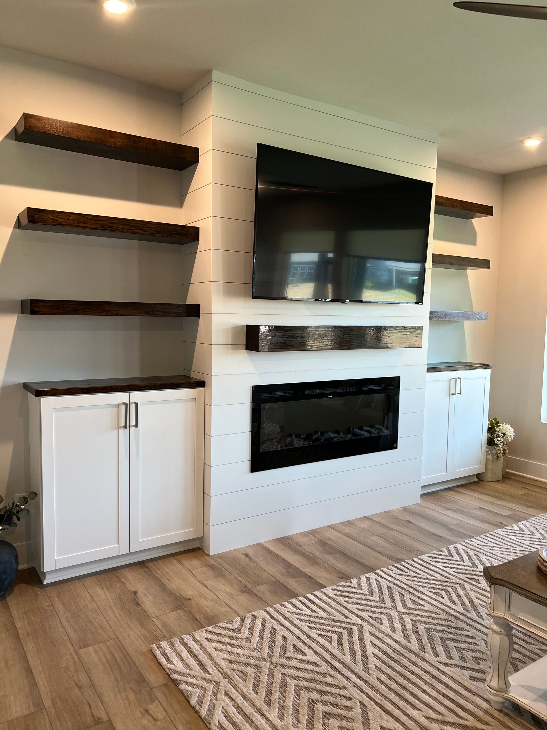 Modern living room wall with mounted TV, electric fireplace, floating shelves, and white cabinets