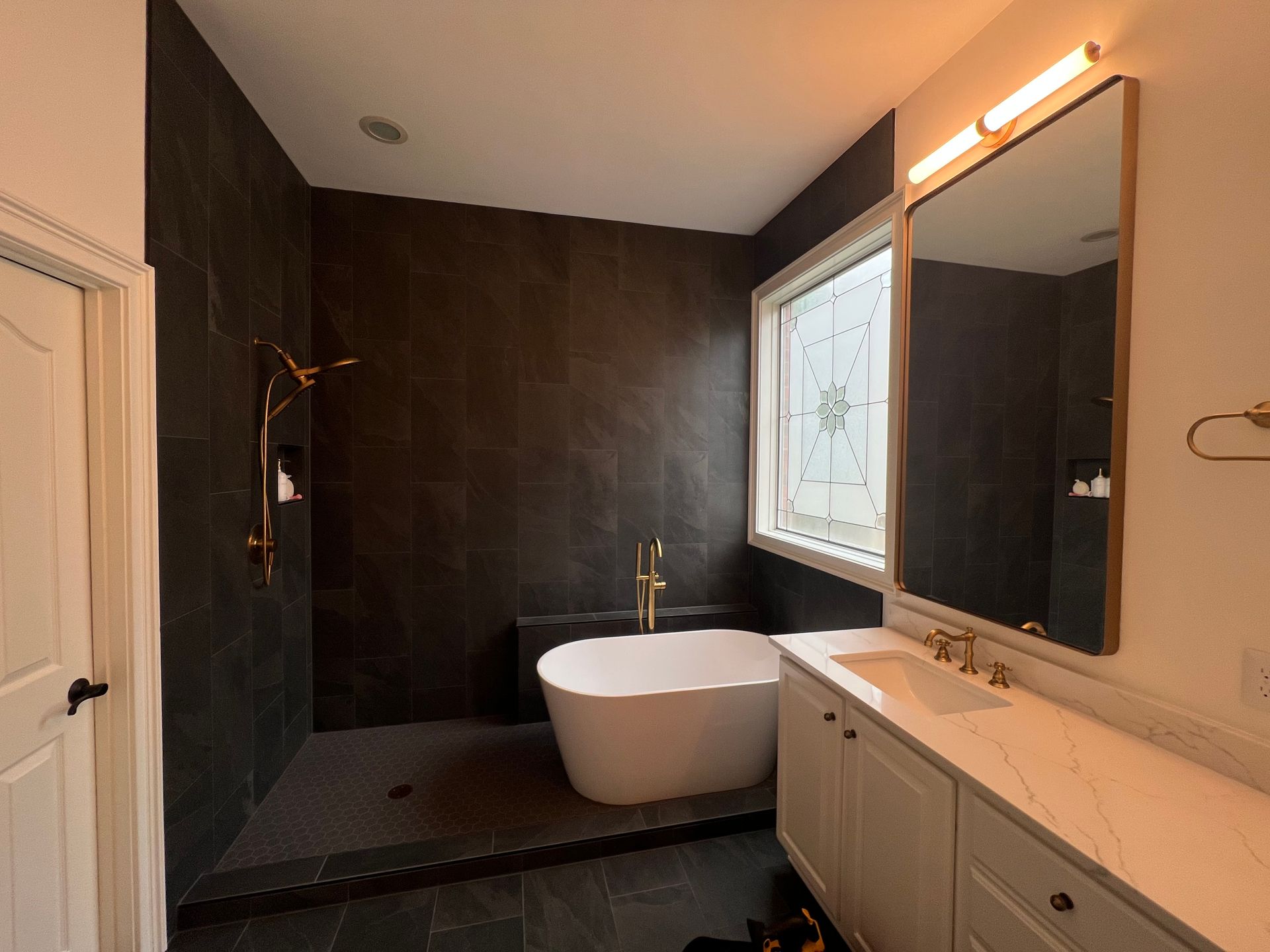Modern bathroom with dark tile shower, freestanding tub, large window, and vanity mirror