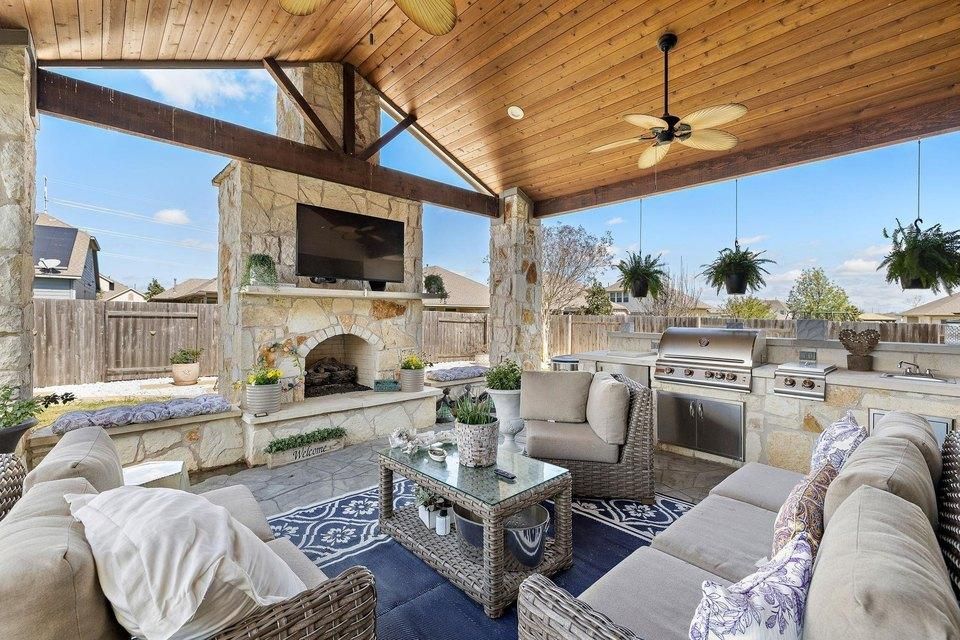 Covered outdoor patio with stone fireplace, sectional seating, and ceiling fan under a wood-paneled roof.