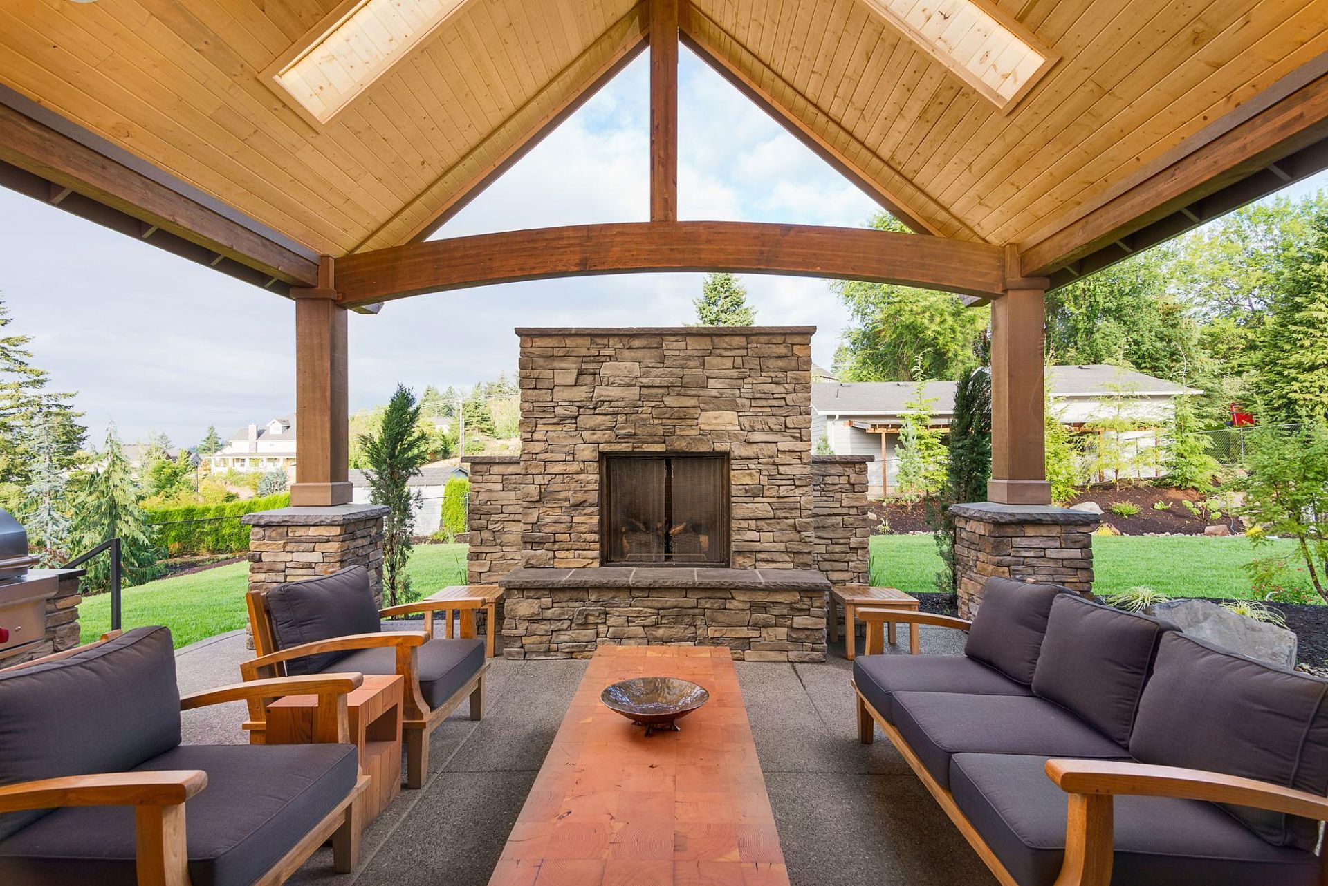 Covered outdoor patio with stone fireplace, wooden ceiling, and cushioned seating around a central table.