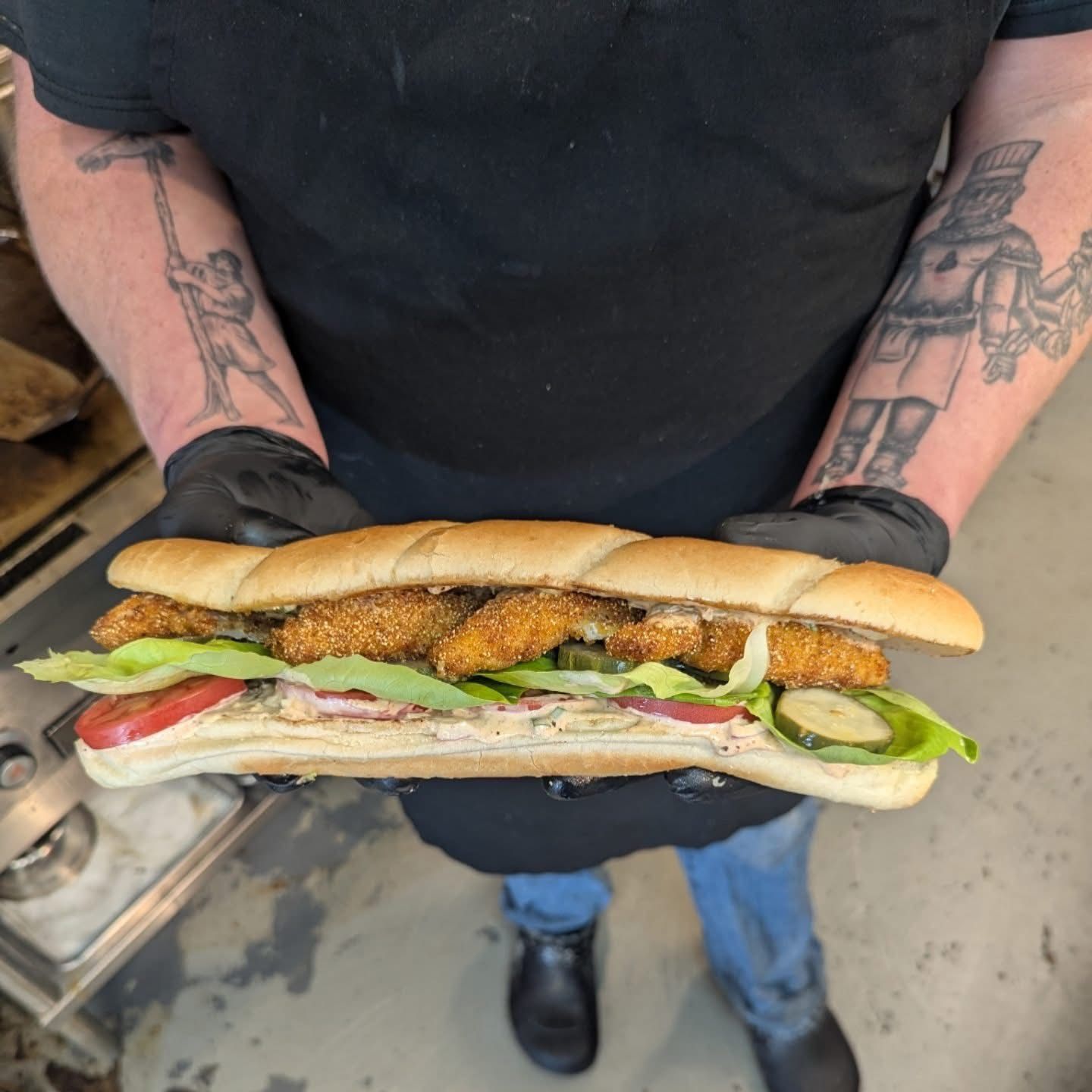 A man wearing black gloves is holding a fried chicken sub sandwich
