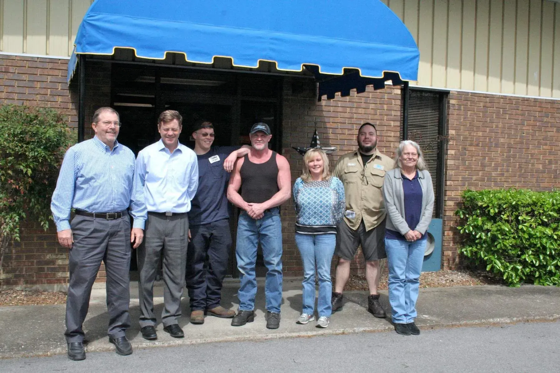 Six people standing outside under a blue awning, posing in front of a brick building