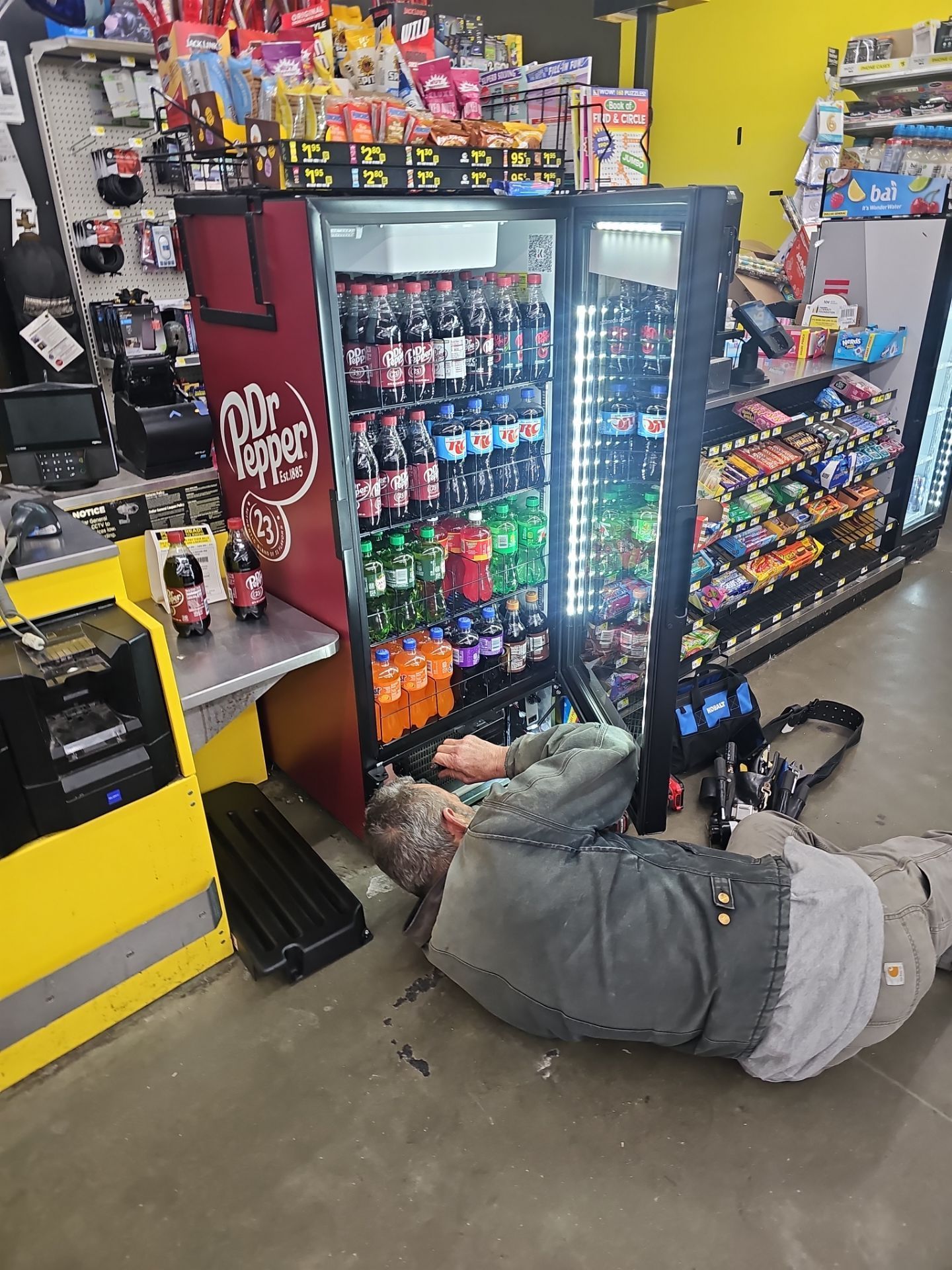 A man is laying on the floor in front of a soda machine in a store.