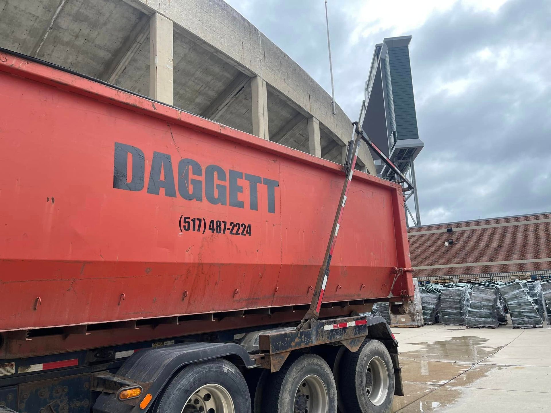 Red DADGET dump trailer at a construction site beside stacked materials and a concrete wall.