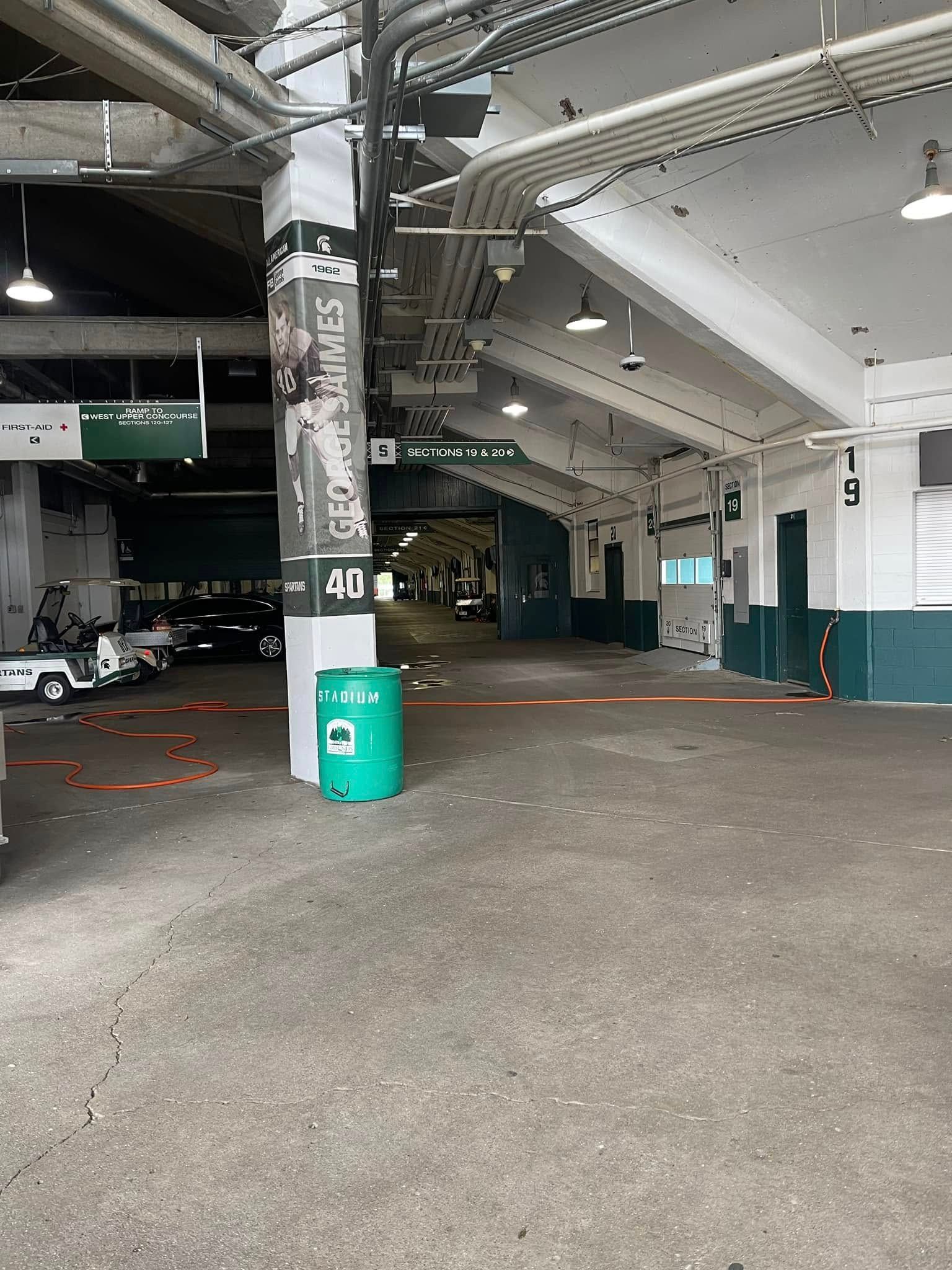Covered parking area with concrete pillars, green trash bin, and parked cars in the background.