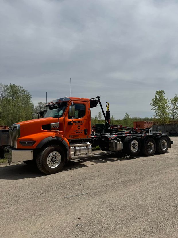 Orange tow truck with flatbed parked on gravel under cloudy sky