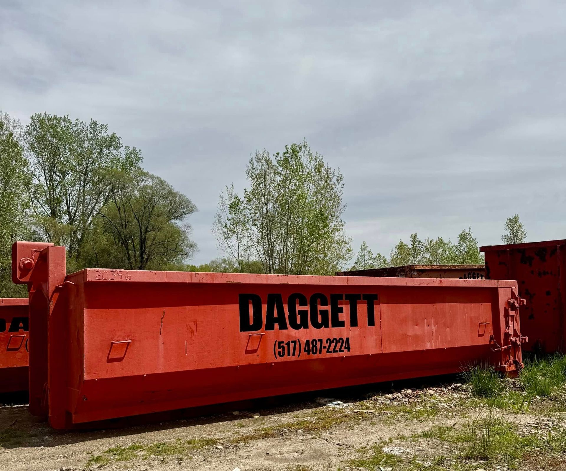 Red dumpster container with “DAGGETT” text parked on gravel under cloudy sky, trees in background