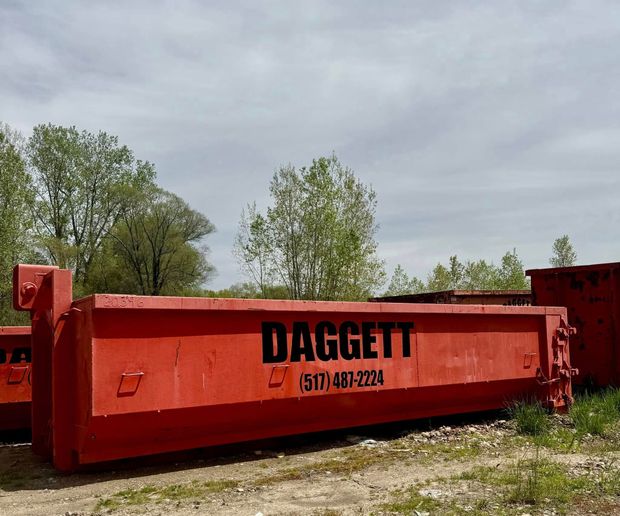 Red metal dumpster marked “DAGGETT” on a gravel lot with trees under a cloudy sky