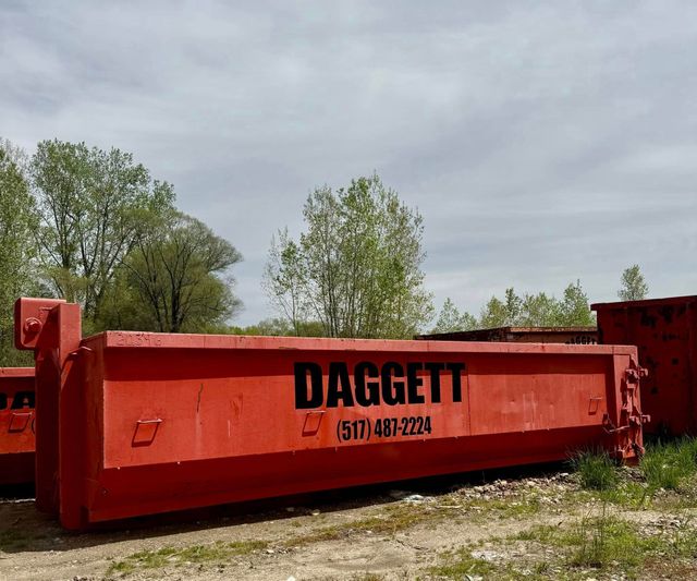 Red metal dumpster marked “DAGGETT” on a gravel lot with trees under a cloudy sky