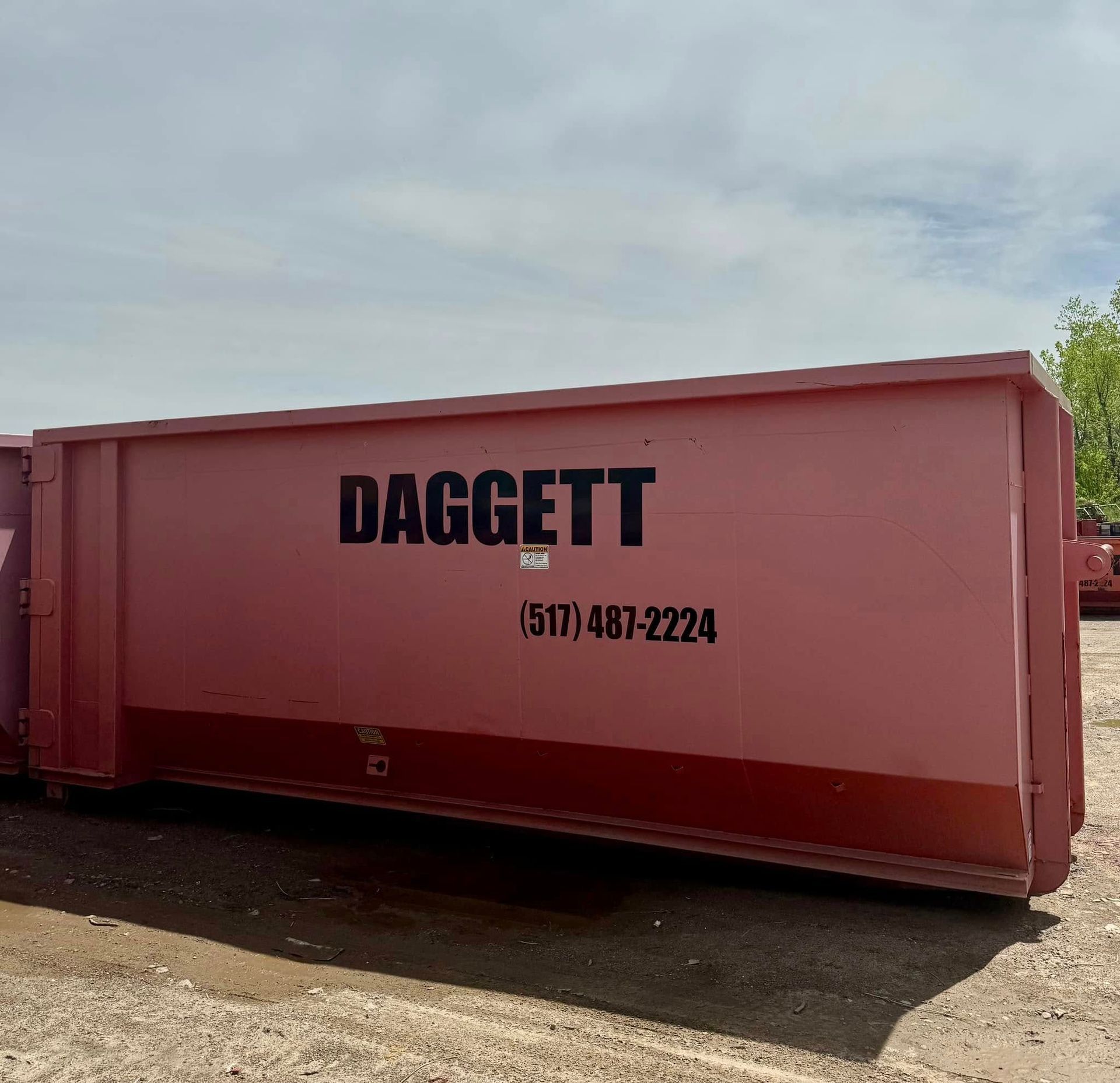 Large red Daggett dumpster container with phone number, outdoors under a cloudy sky