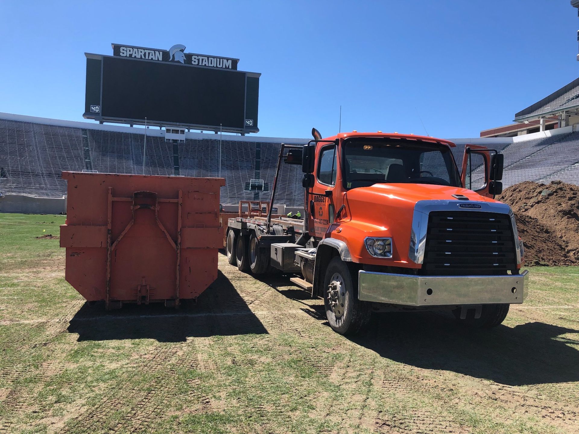 Orange truck and red dumpster on a grassy stadium field with empty bleachers and scoreboard in the background