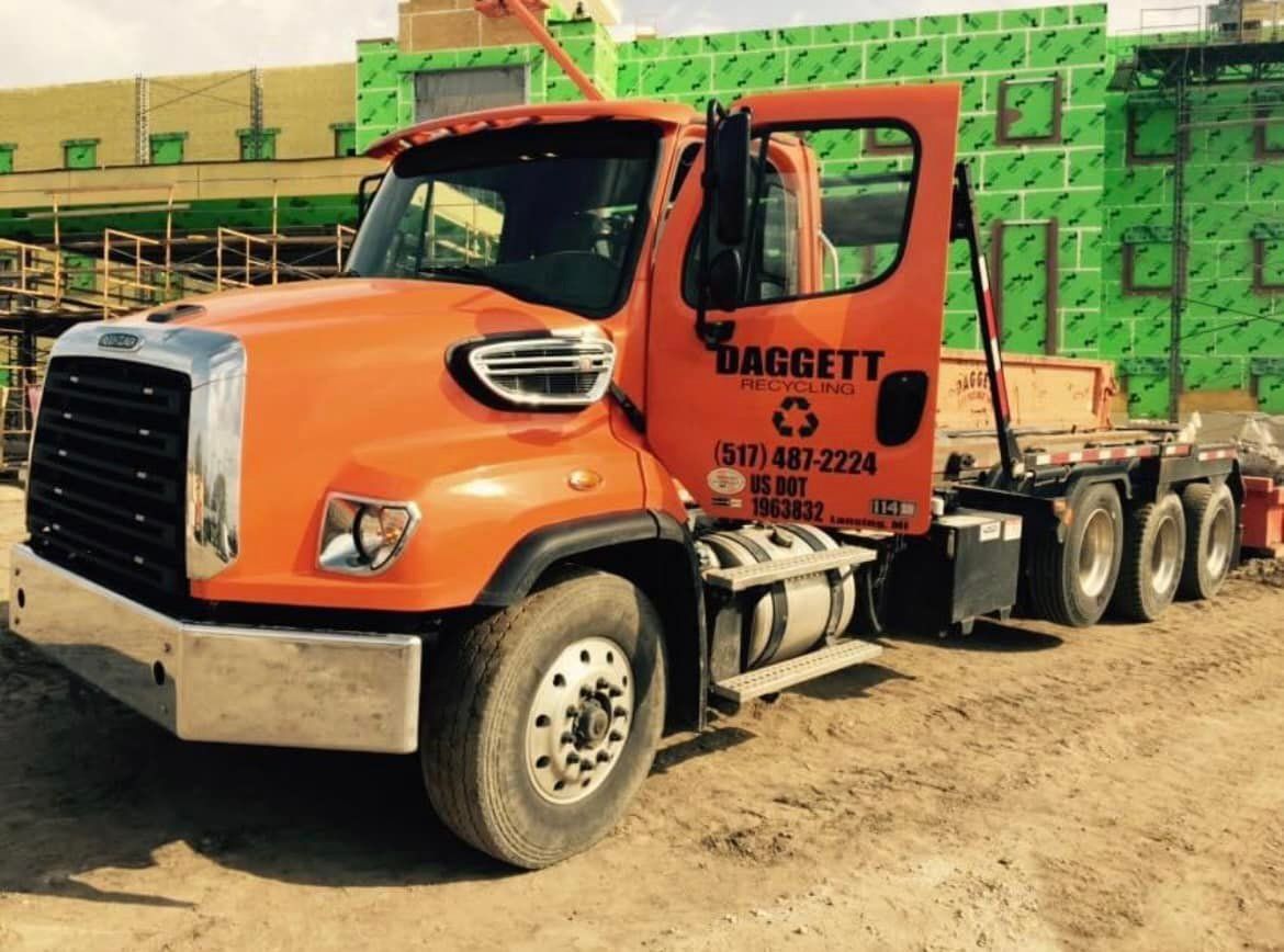 Orange semi-truck with trailer parked at a construction site beside green building materials.