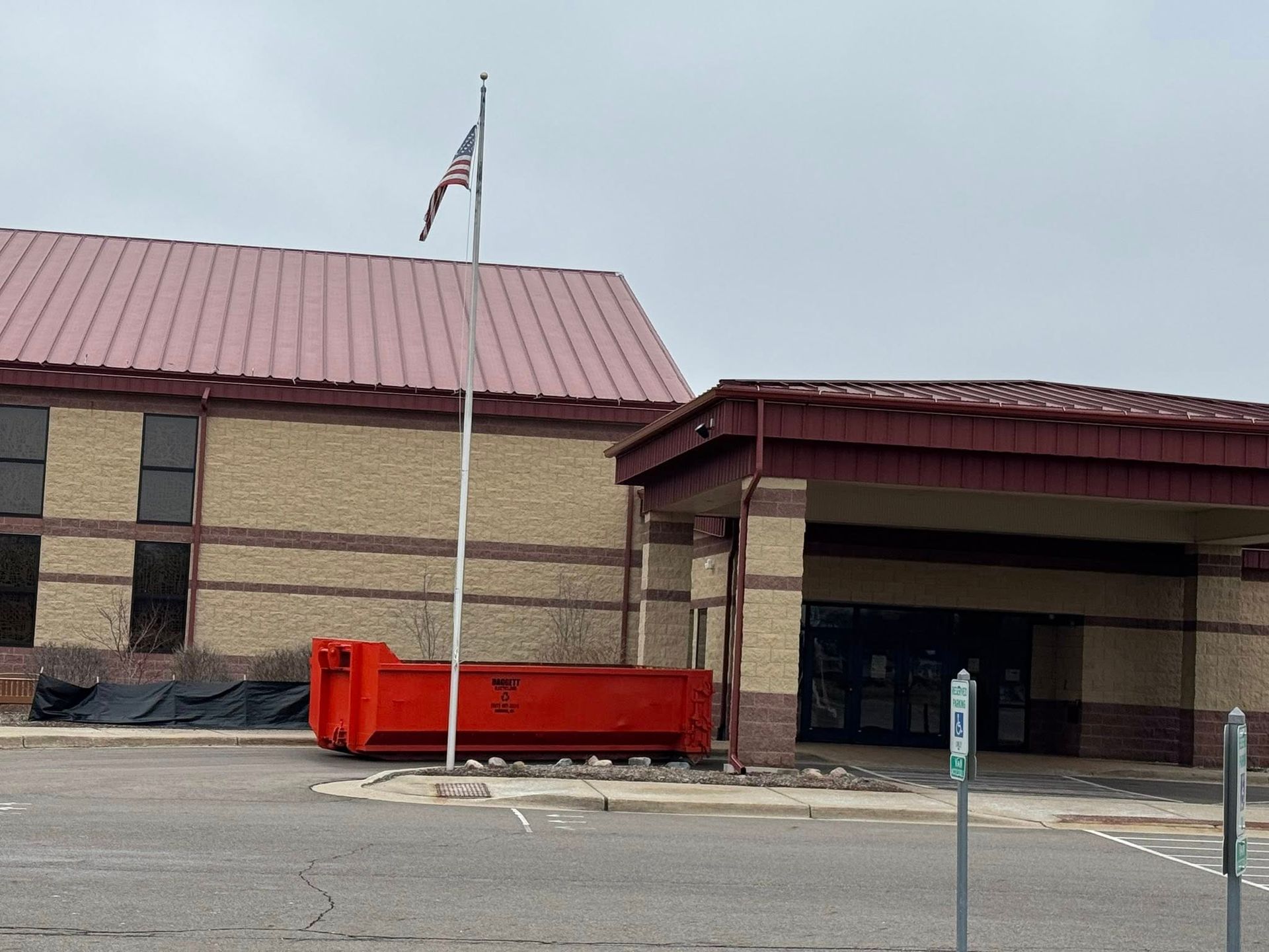 Low brick building with a red roof, flagpole, and red barricade in front under an overcast sky