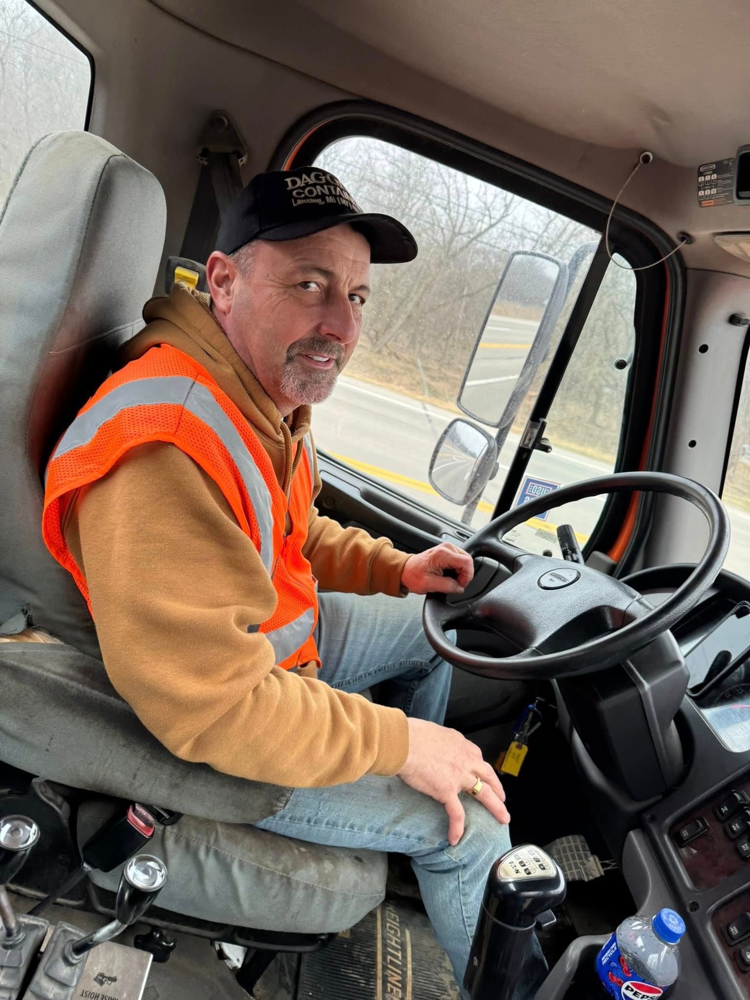 Worker in orange safety vest sitting in a truck cab, holding the steering wheel.