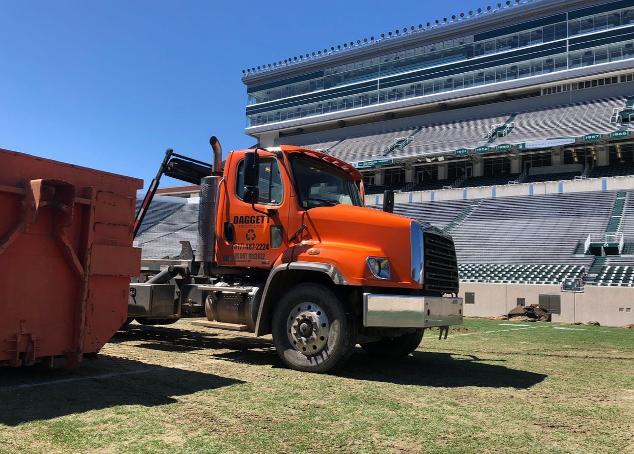 Orange tow truck beside a red dumpster in a stadium under clear blue sky