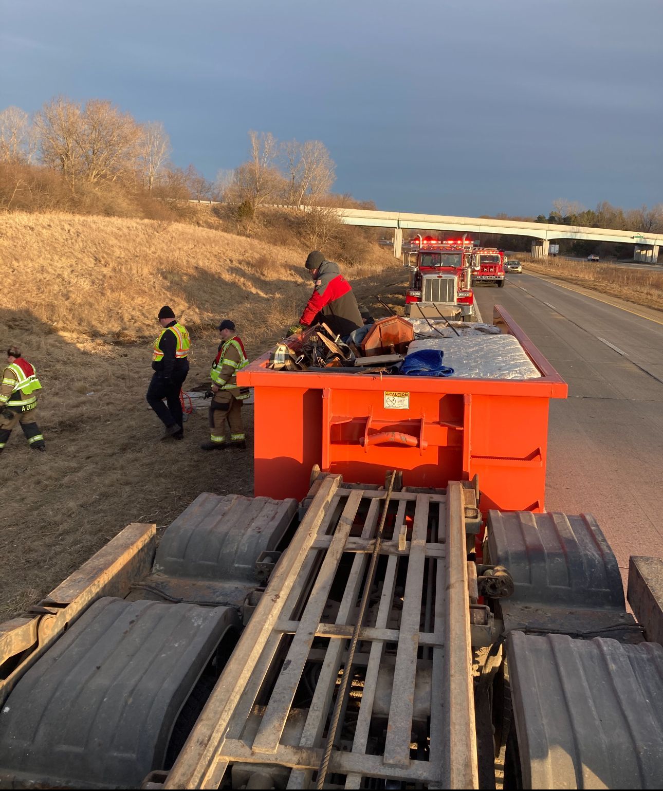Rescue workers beside an overturned orange truck on a highway shoulder after a crash