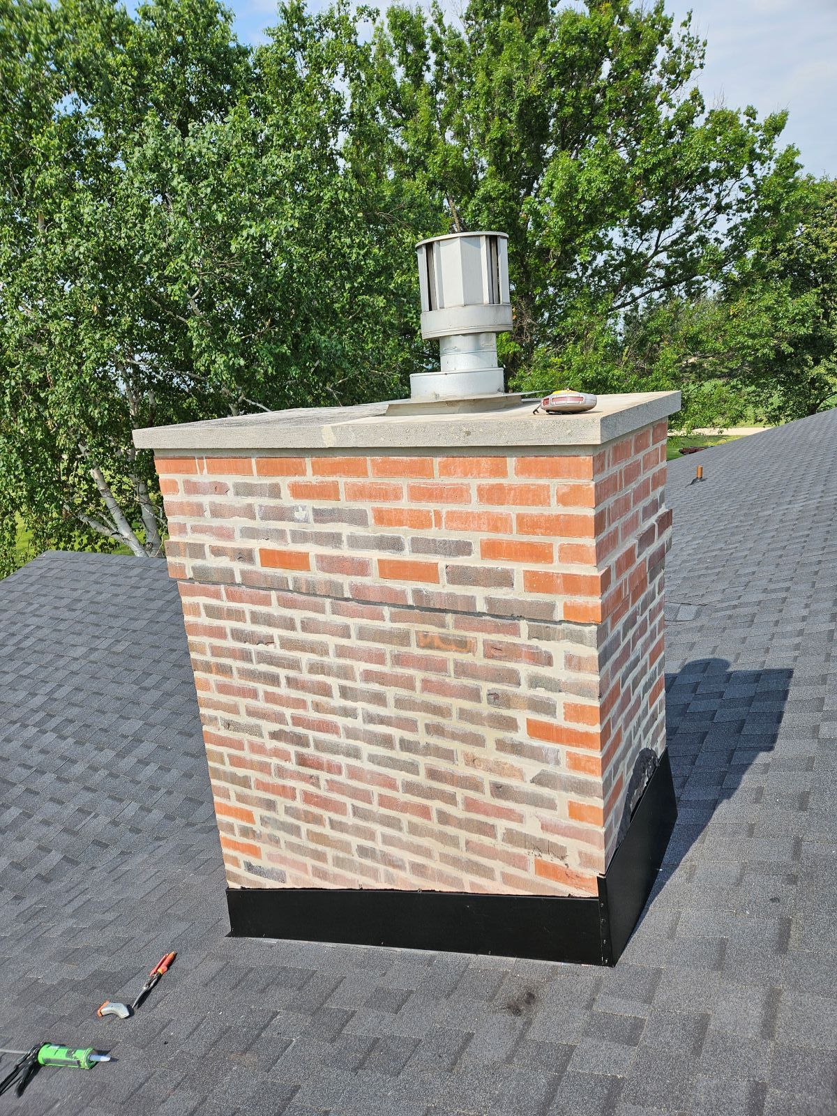 Brick chimney with metal cap on a black shingled roof, trees in the background.