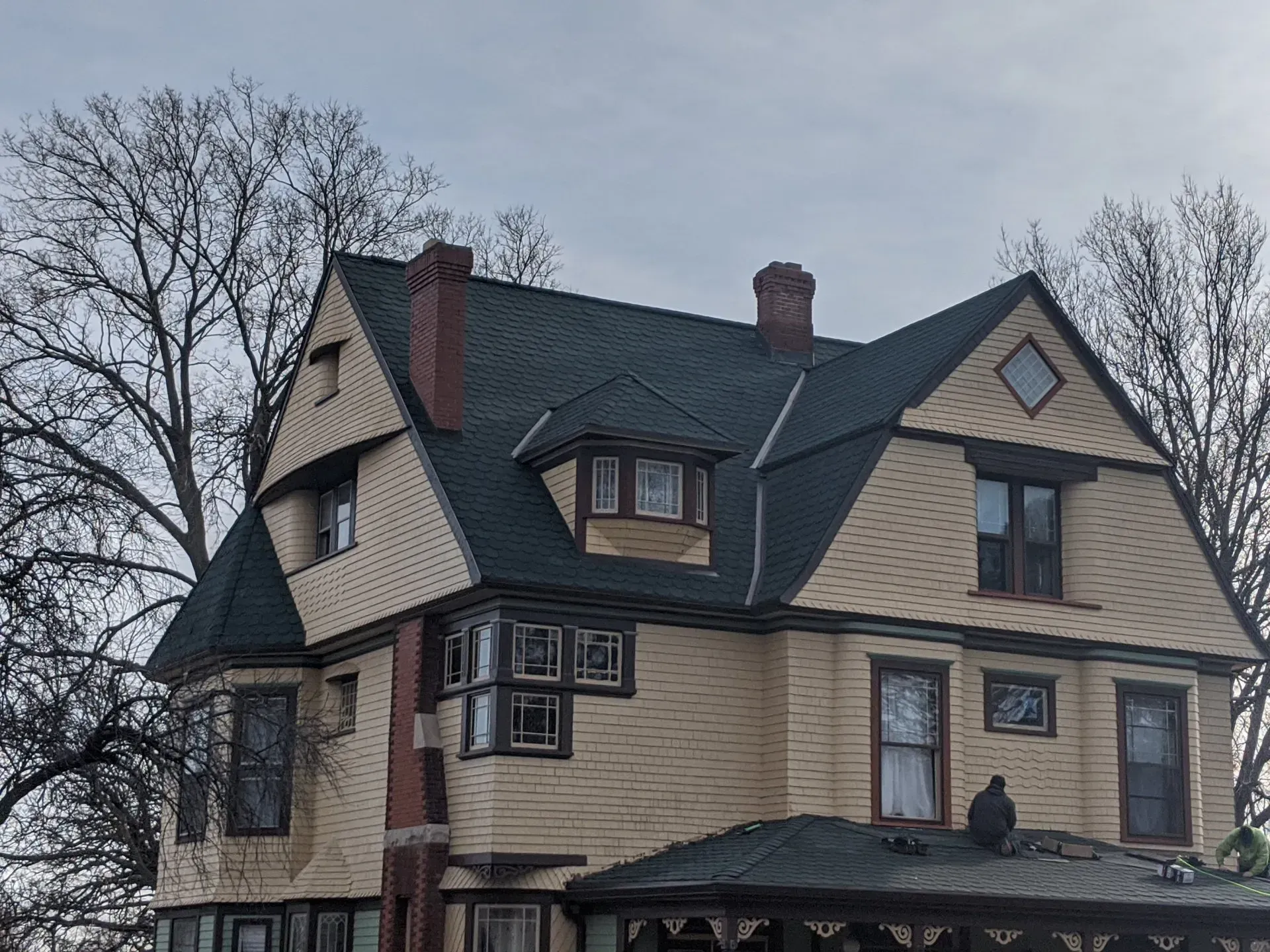 Yellow house with dark green roof, multiple gables, windows, and two brick chimneys against a cloudy sky.