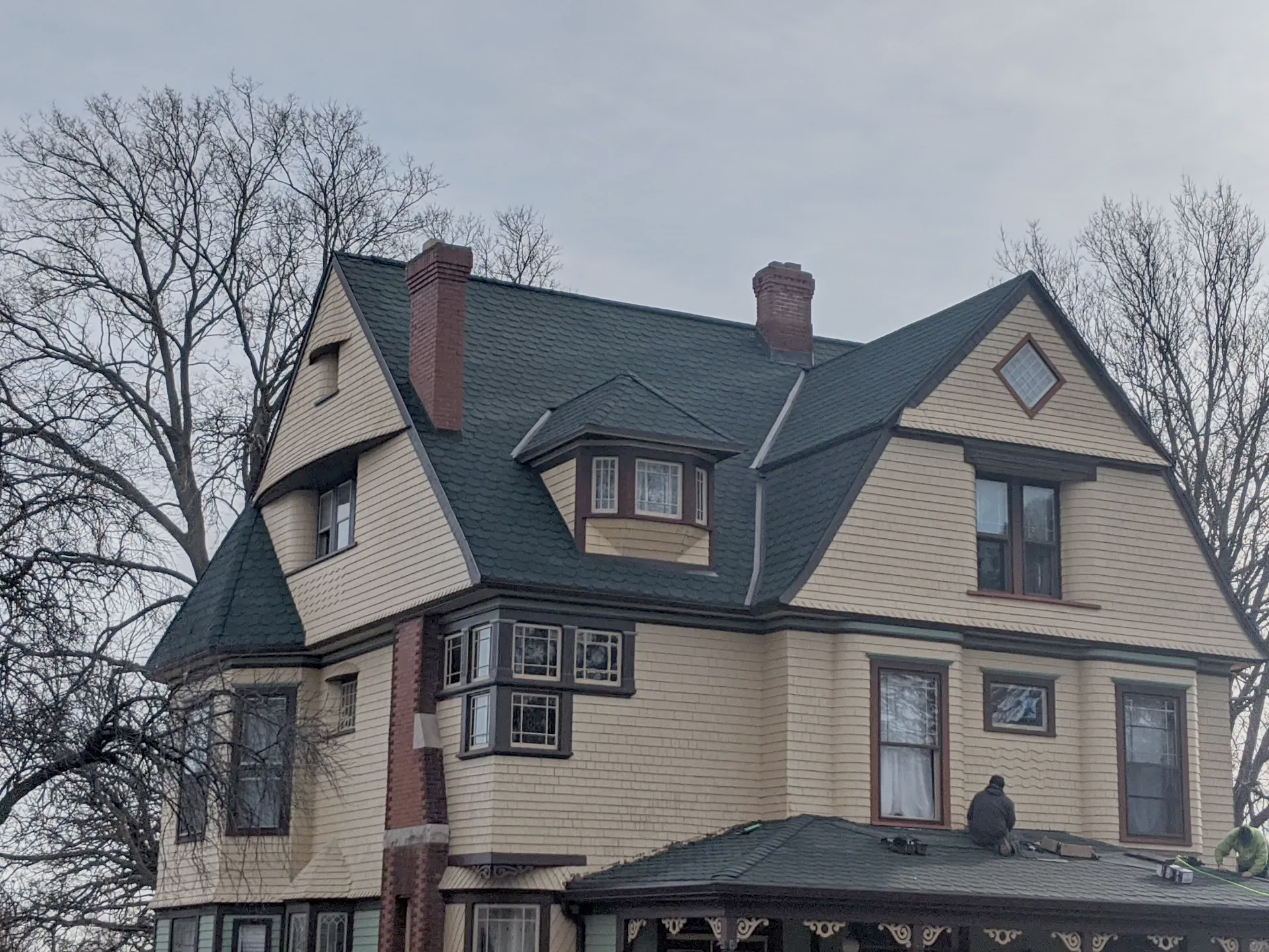 Yellow house with dark green roof, multiple gables, windows, and two brick chimneys against a cloudy sky.