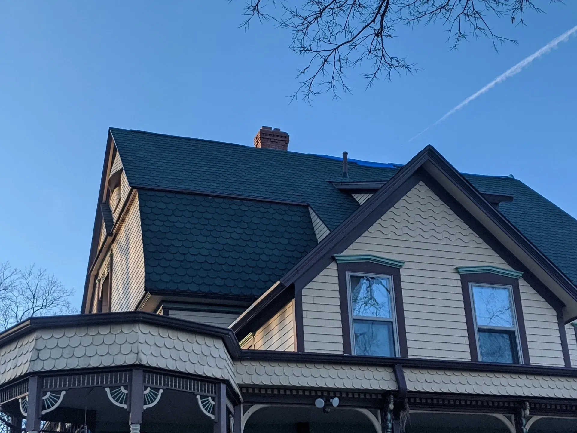 Victorian house with dark green roof and blue tarp against a blue sky, chimney visible.