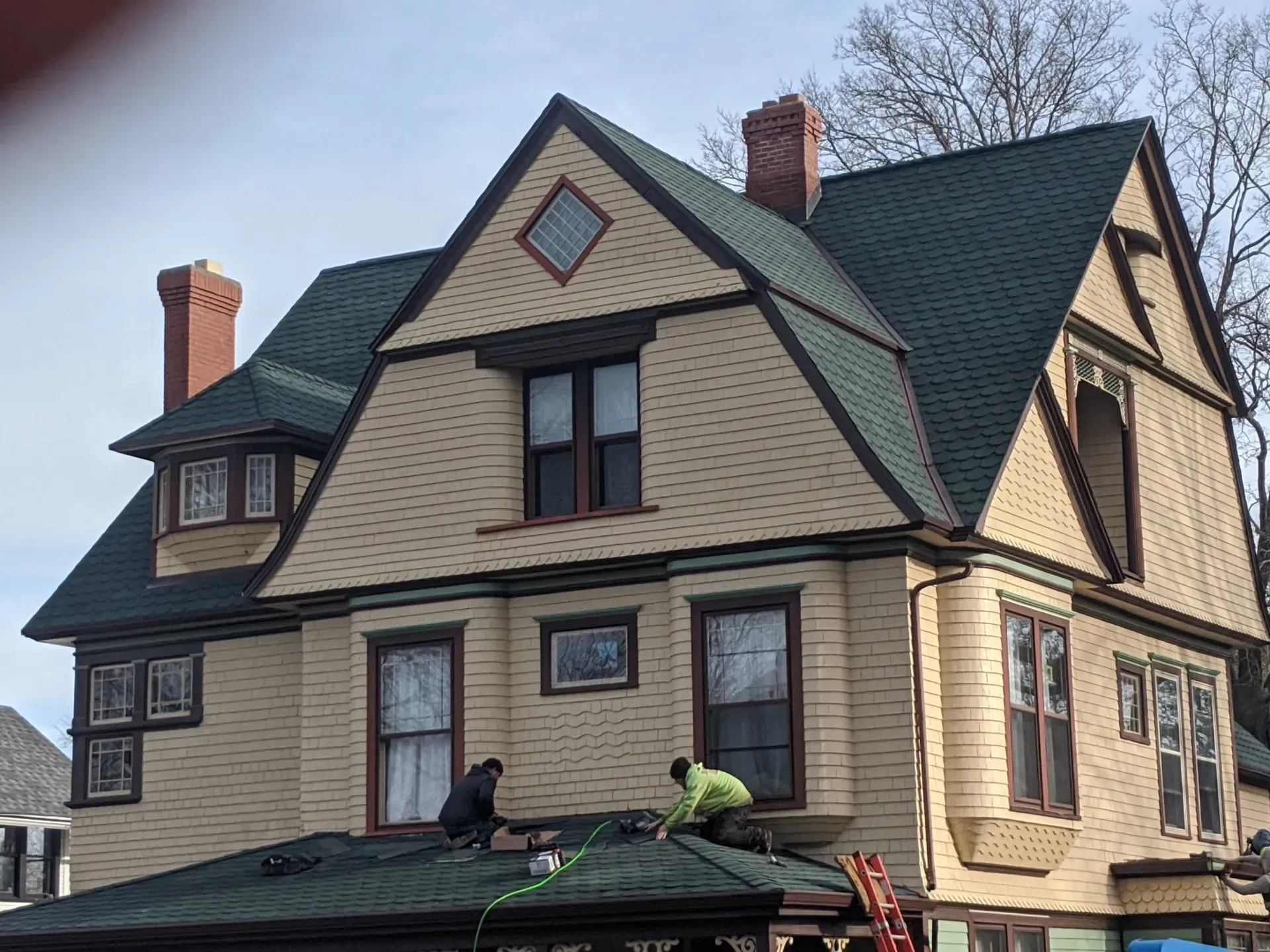 Two workers on a green-roofed house with tan siding and brown trim. Red brick chimney.