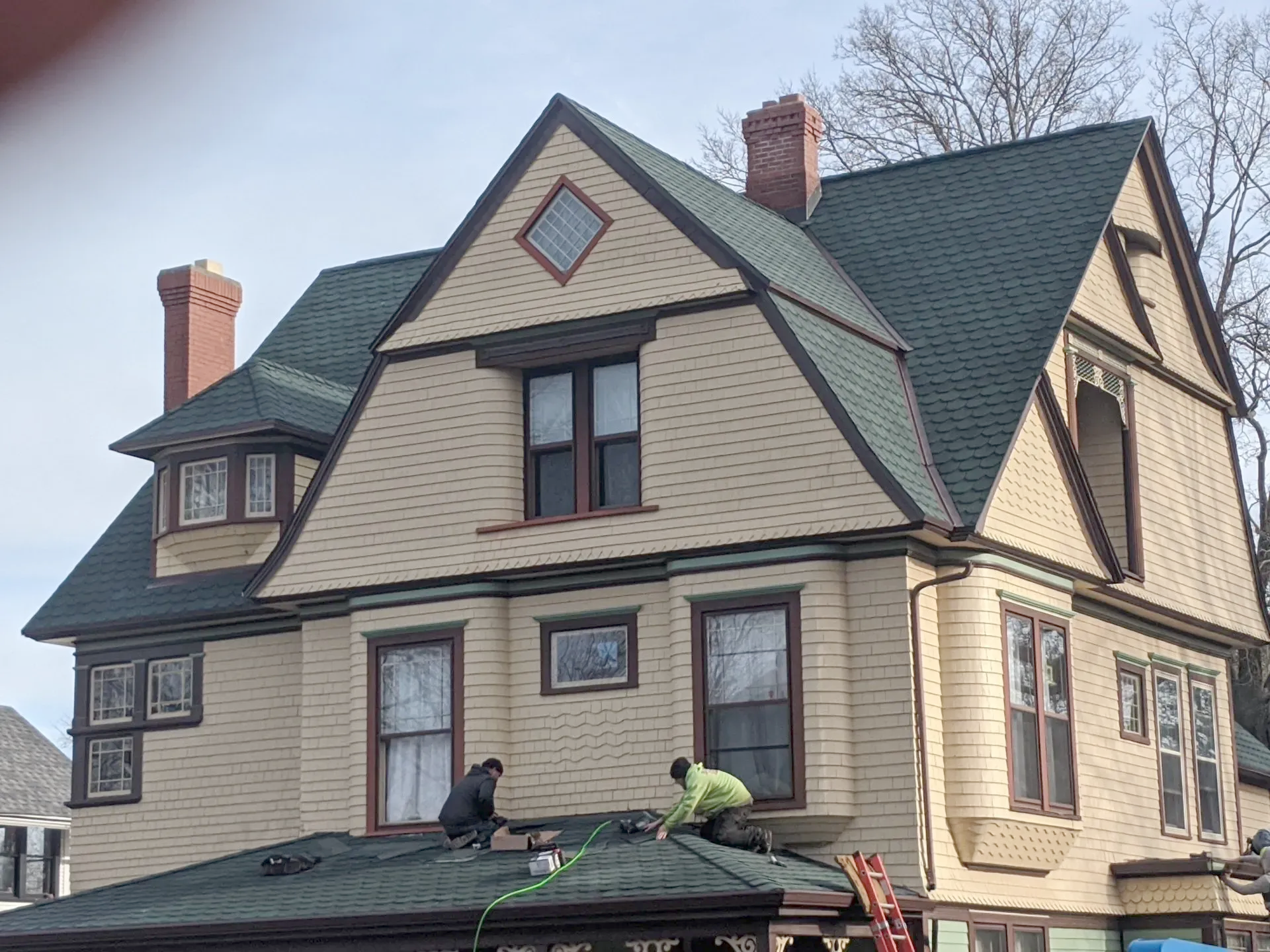 Workers on a roof of a beige Victorian house with green roofing and trim, under a clear sky.