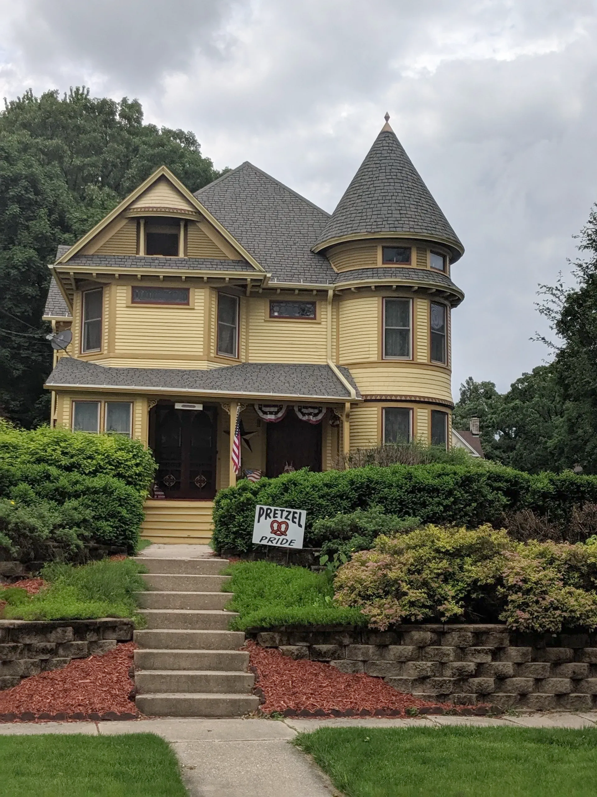 Yellow Victorian house with a turret, steps, and front yard landscaping. Cloudy sky.