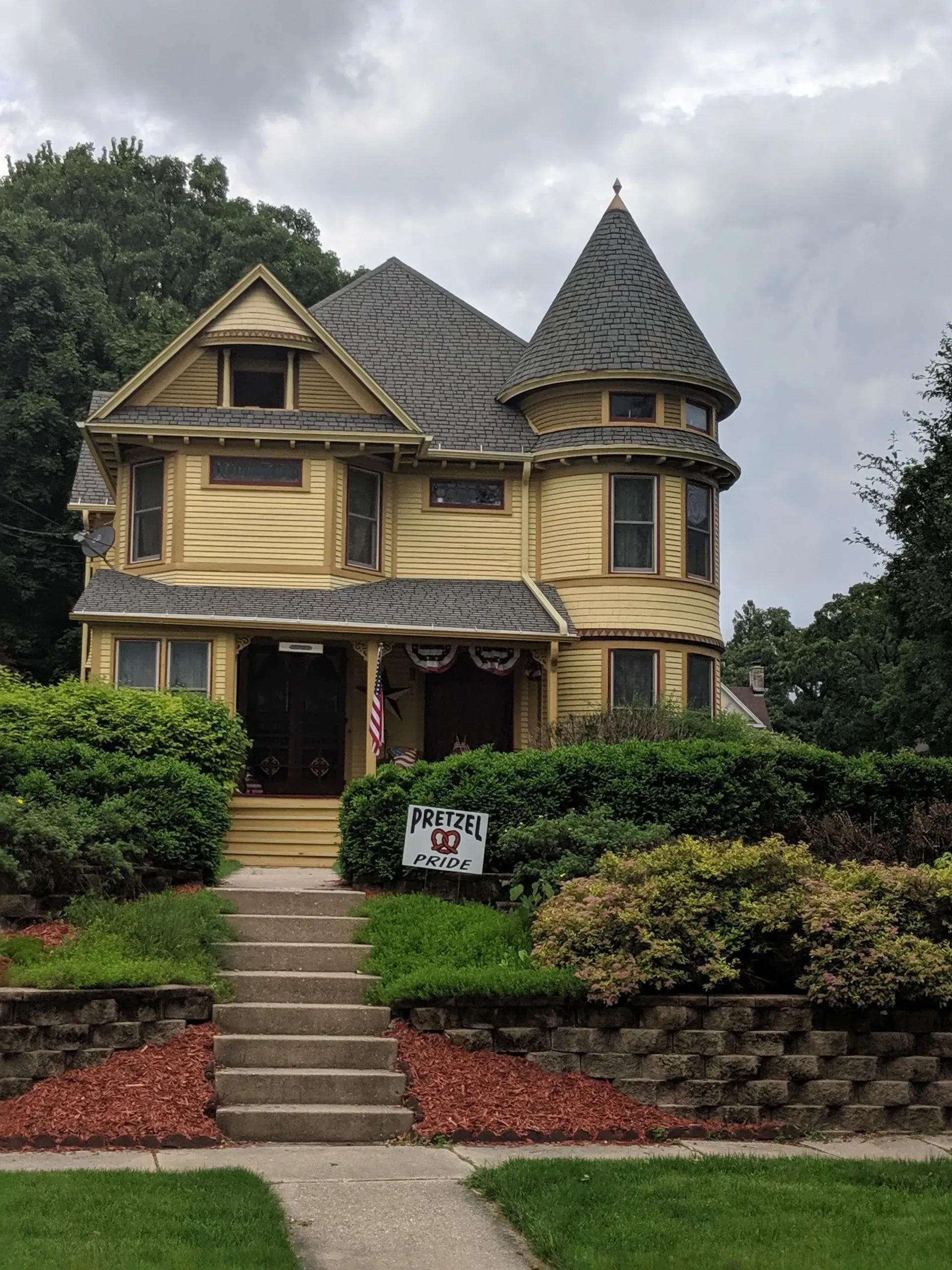 Yellow Victorian house with a turret, steps, and front yard landscaping. Cloudy sky.