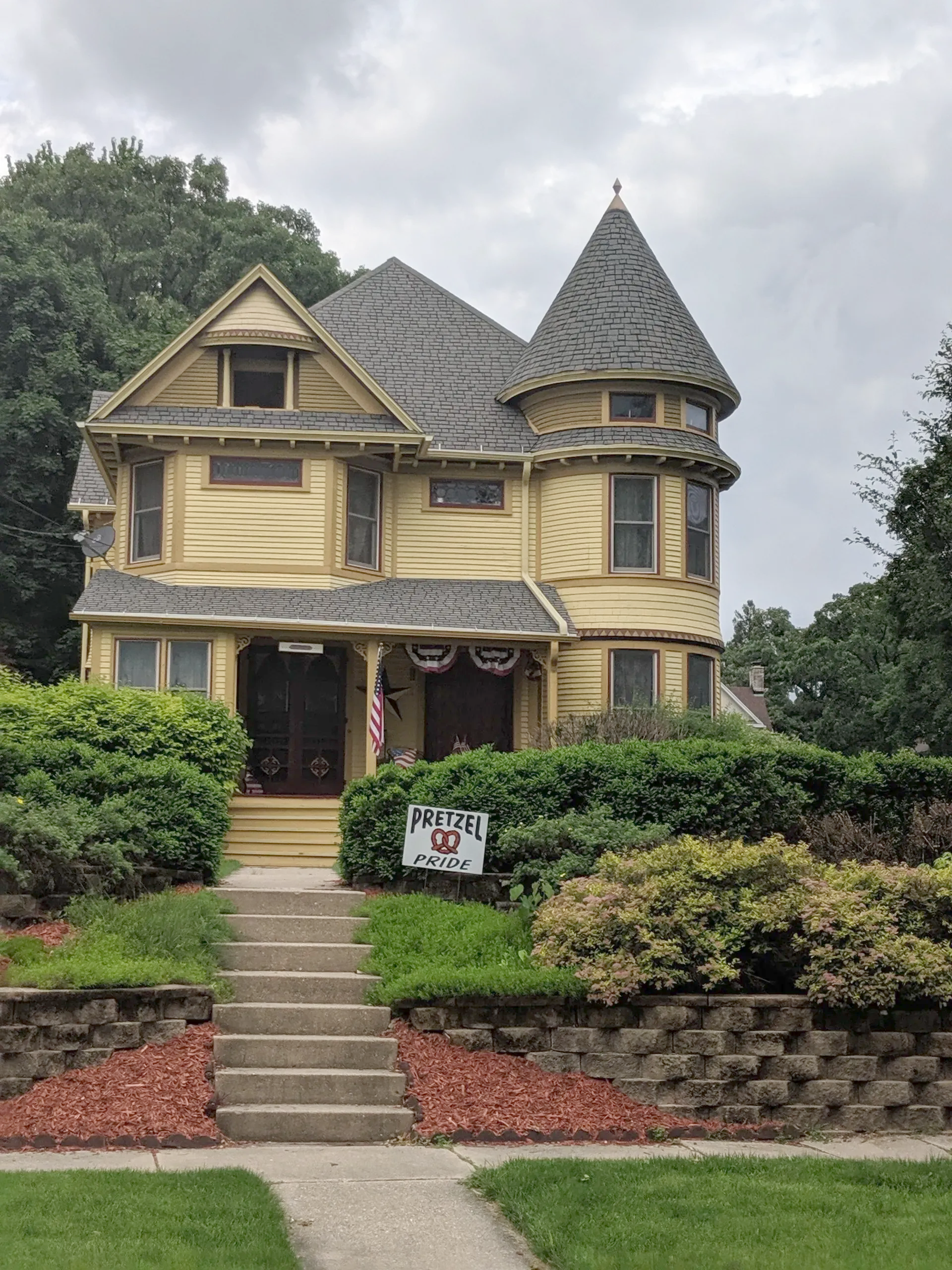 Yellow Victorian house with a turret, steps, and front yard landscaping under a cloudy sky.