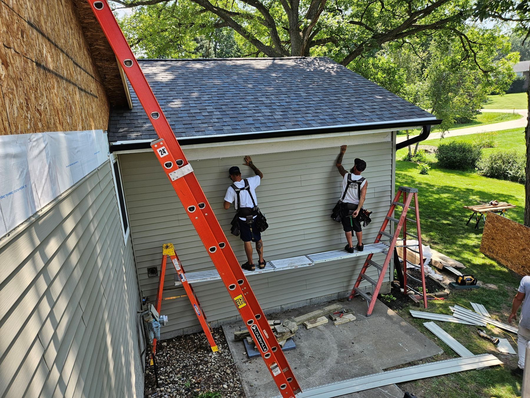 Two people install siding on a building's exterior. One works from a platform, the other from a ladder.