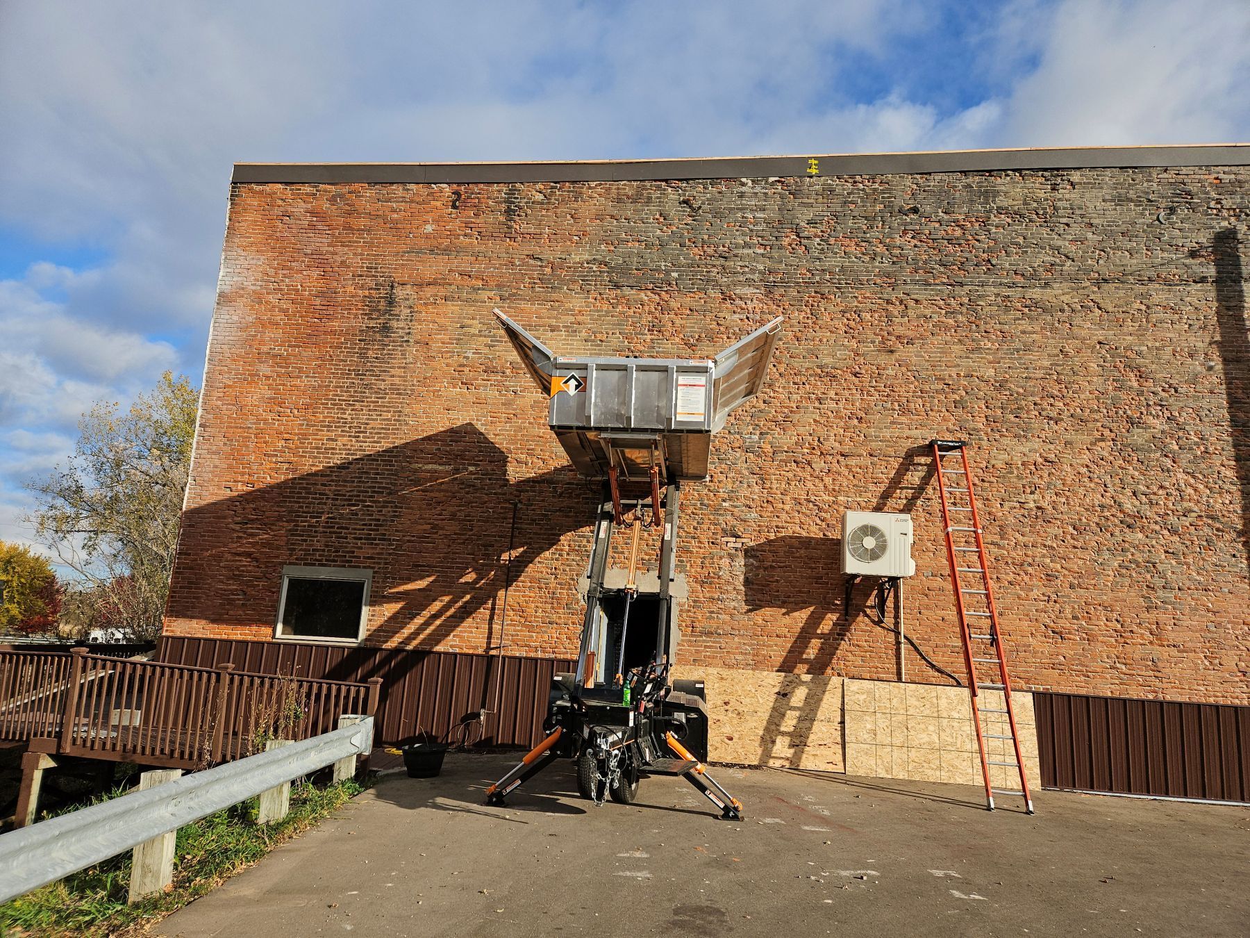 A lift platform against a brick building, loading materials. A ladder and air conditioner are on the wall.