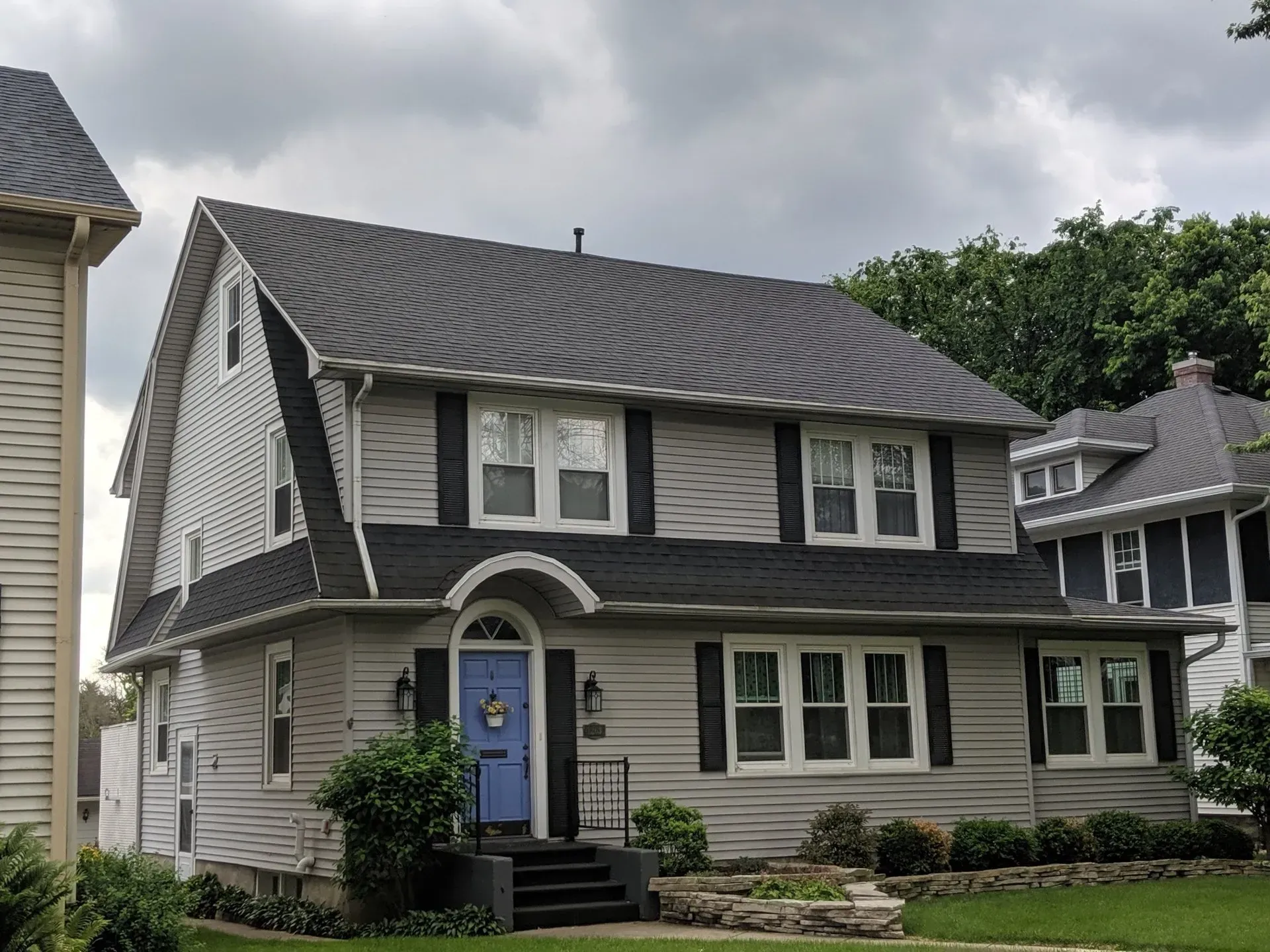 Two-story house with gray siding, black shutters, blue front door, and a dark roof under a cloudy sky.