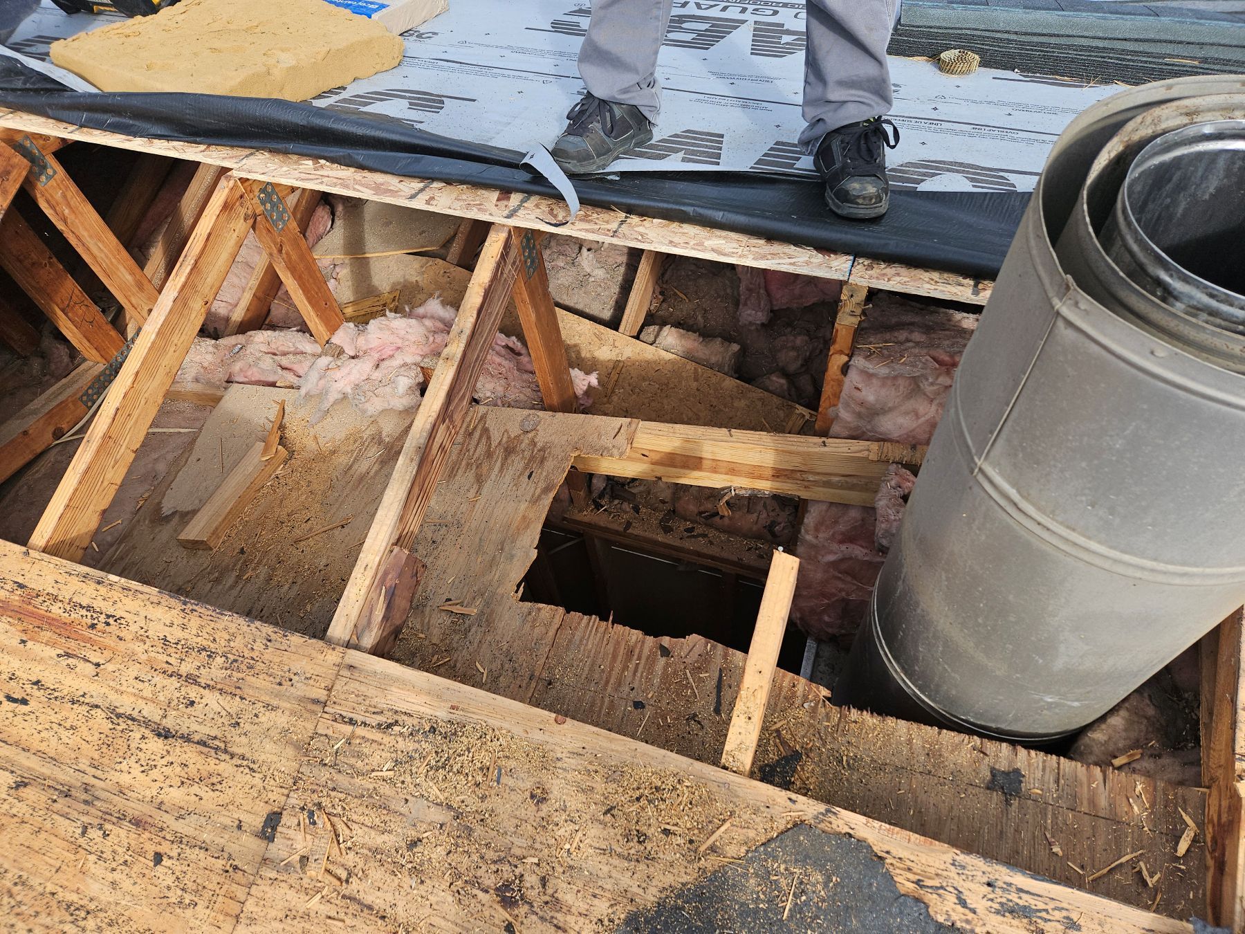Man standing on exposed roof structure, holding metal chimney pipe. Wood frame with insulation visible.