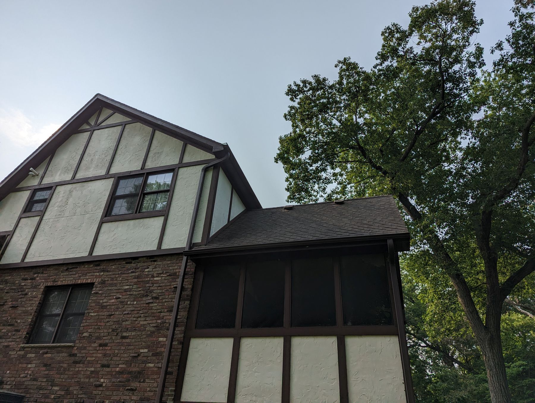 Two-story house with brown and white exterior, screened porch, and large green tree.