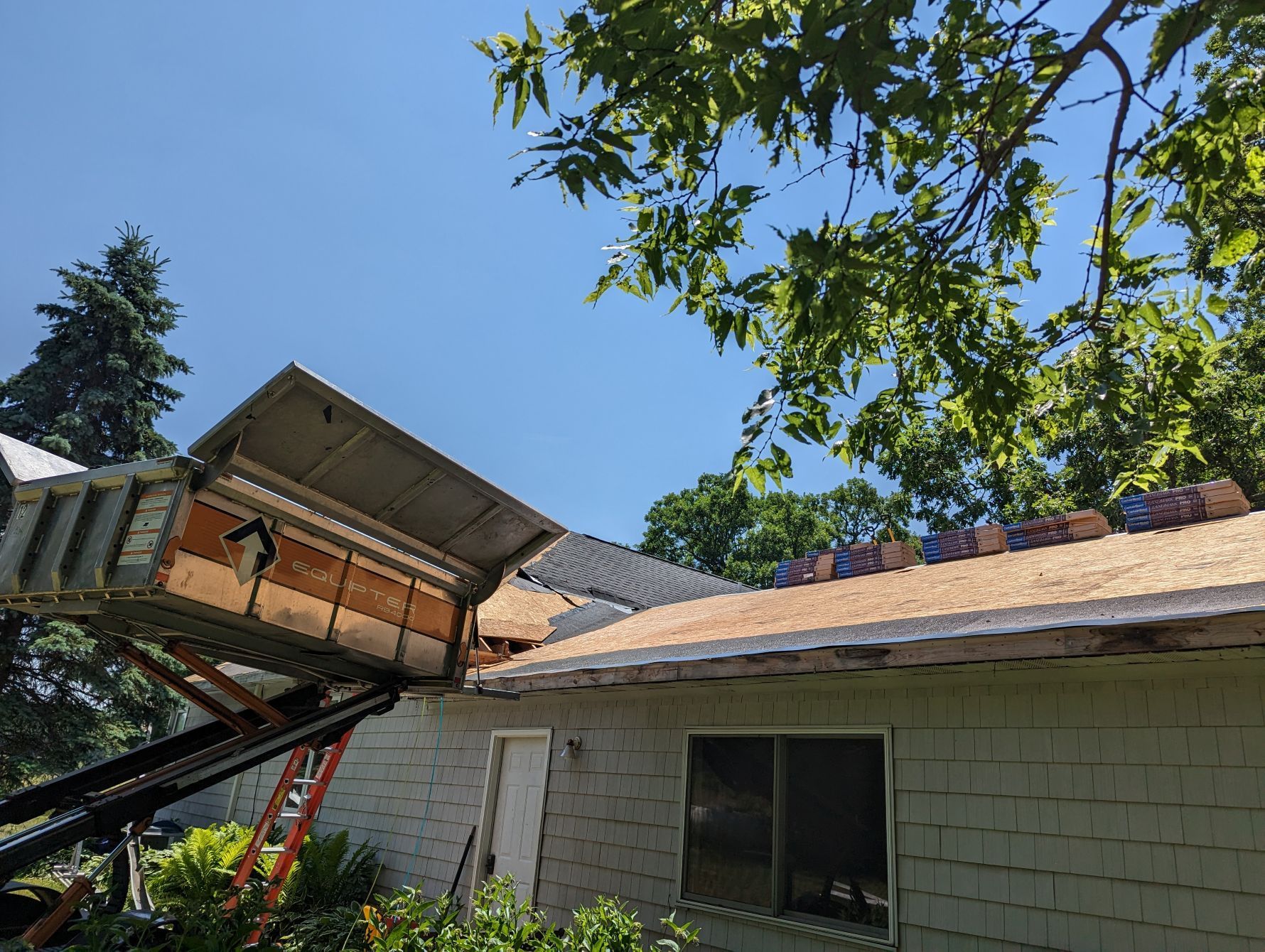 House roof being repaired with debris chute in use; blue sky, green trees, sun.