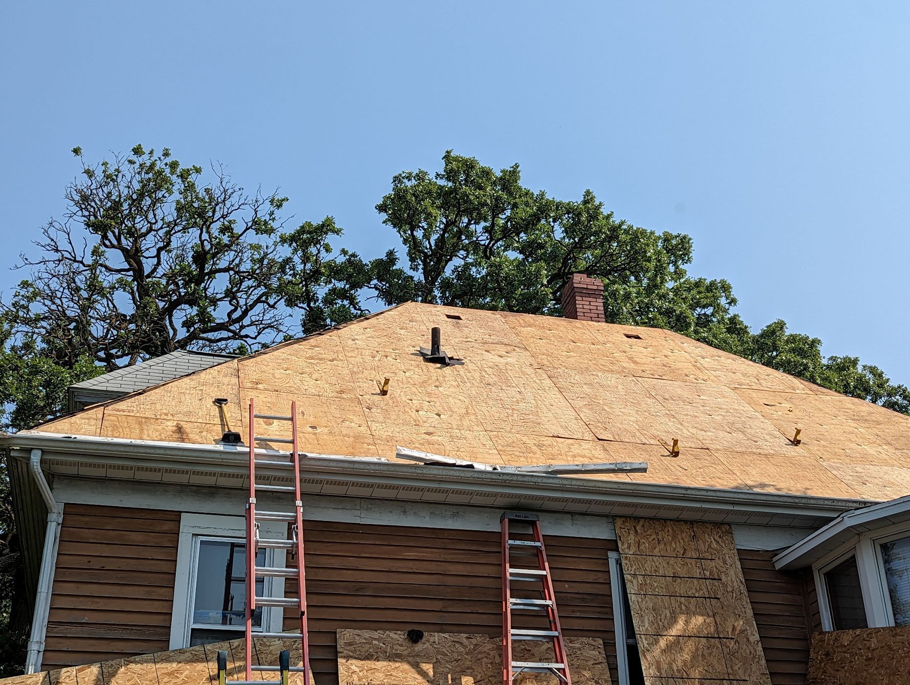 House roof under construction, plywood installed. Two ladders lean against the building. Bright, sunny day.