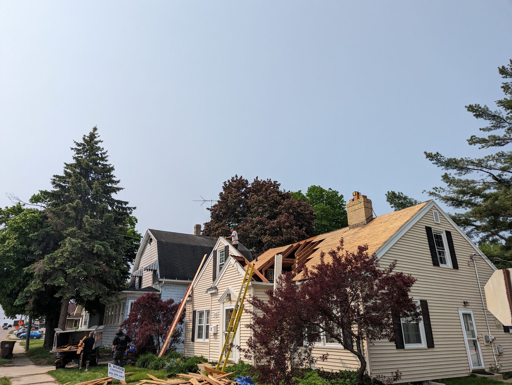 House with roof partially removed, construction underway, surrounded by trees and other houses.