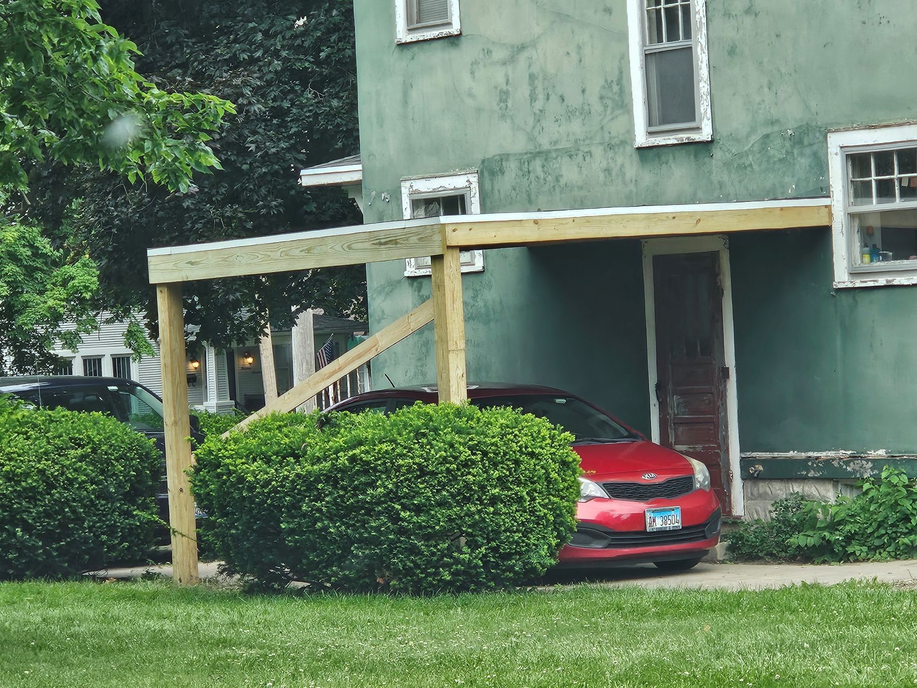 Red car parked under a wooden carport attached to a green building.