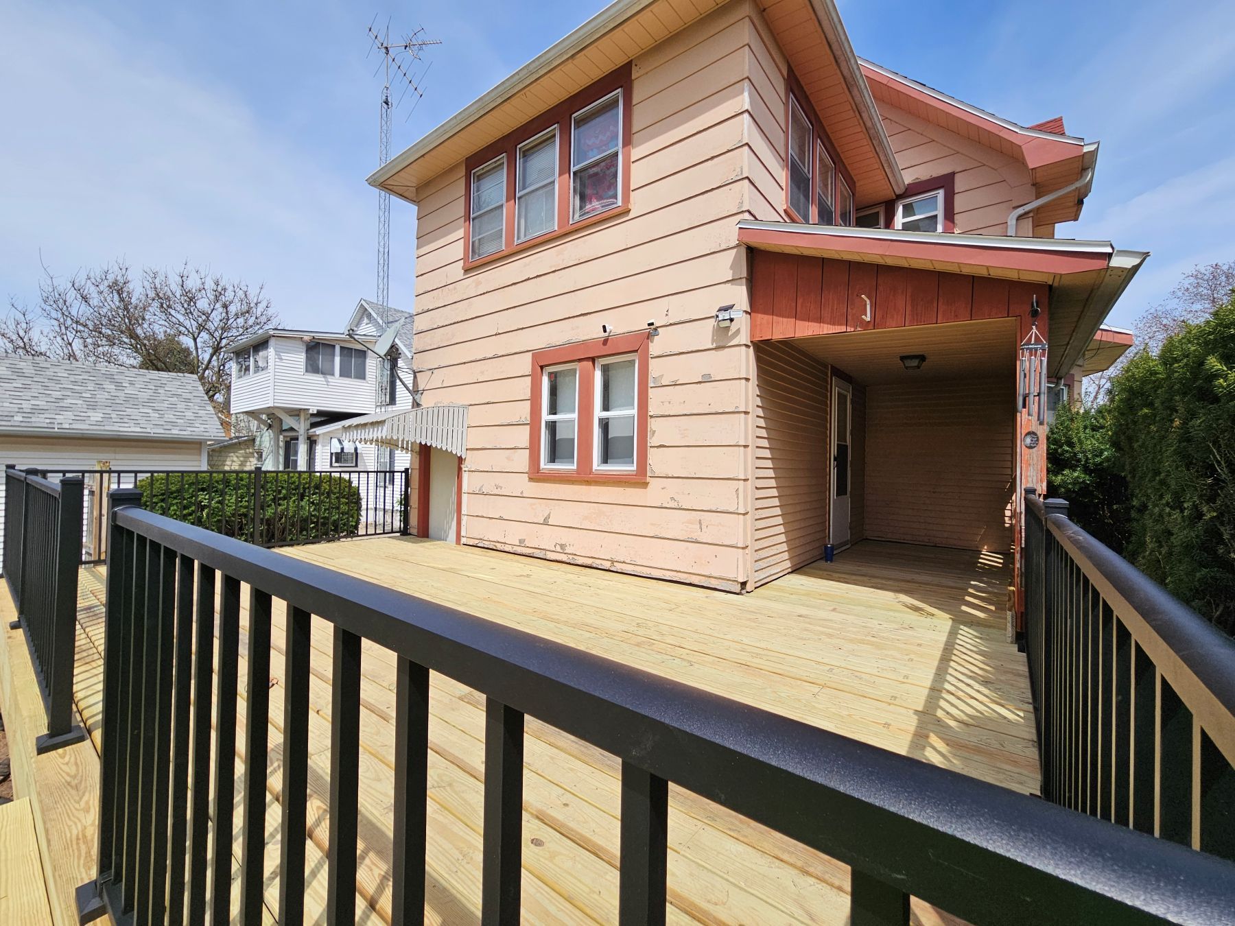 Exterior view of a two-story house with a wooden deck and black railings under a blue sky.