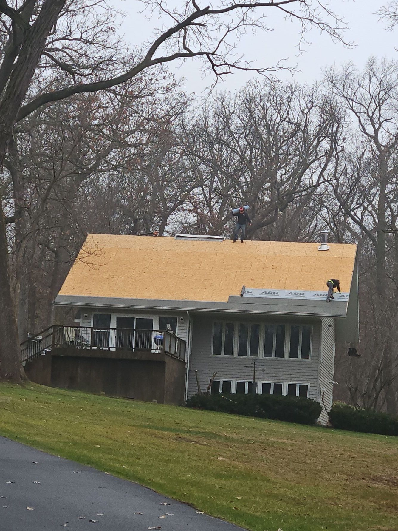 Workers installing roofing on a house with large windows, surrounded by trees on a cloudy day.