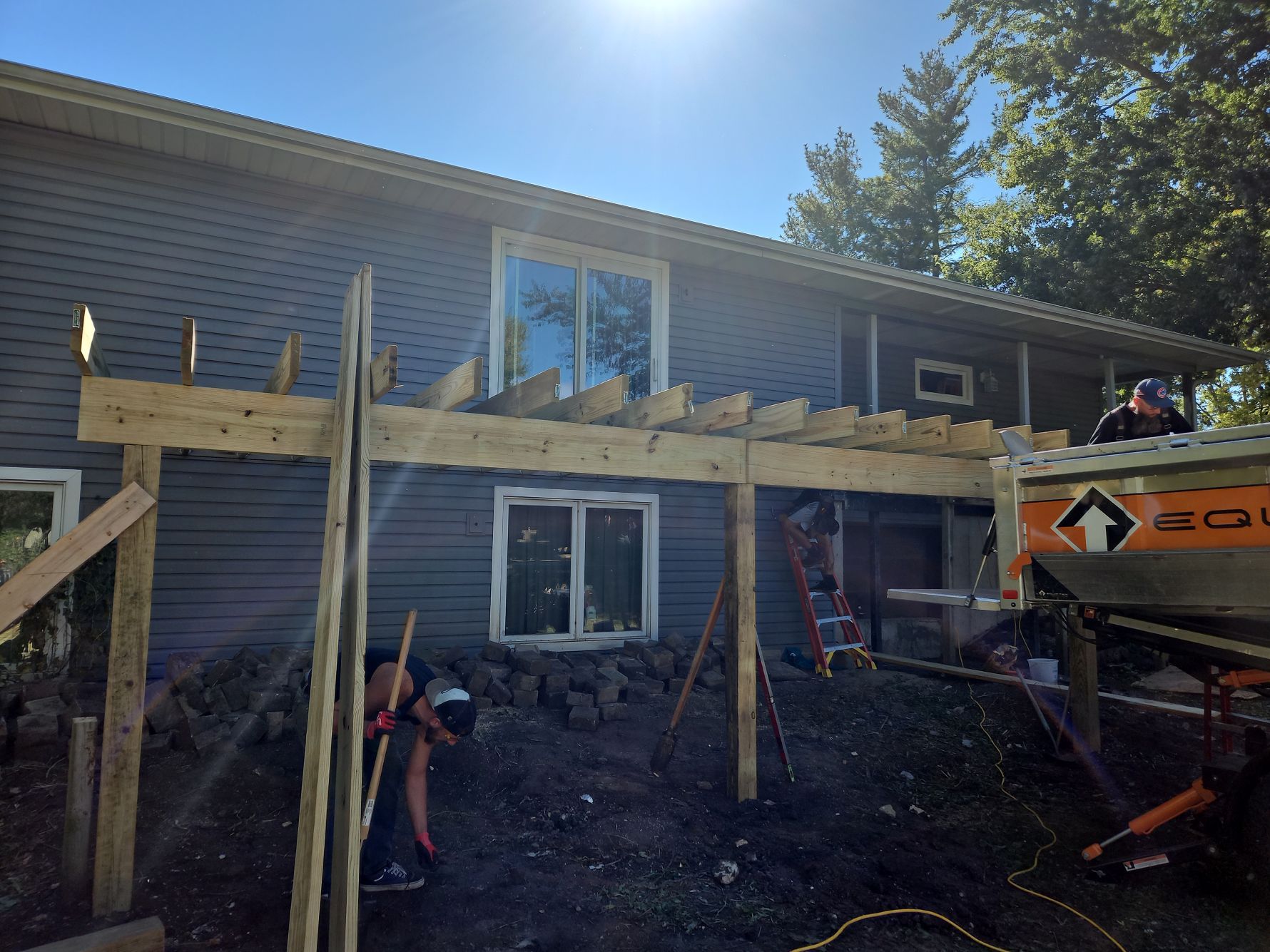 Construction of a wooden deck attached to a gray house, with workers and materials visible.