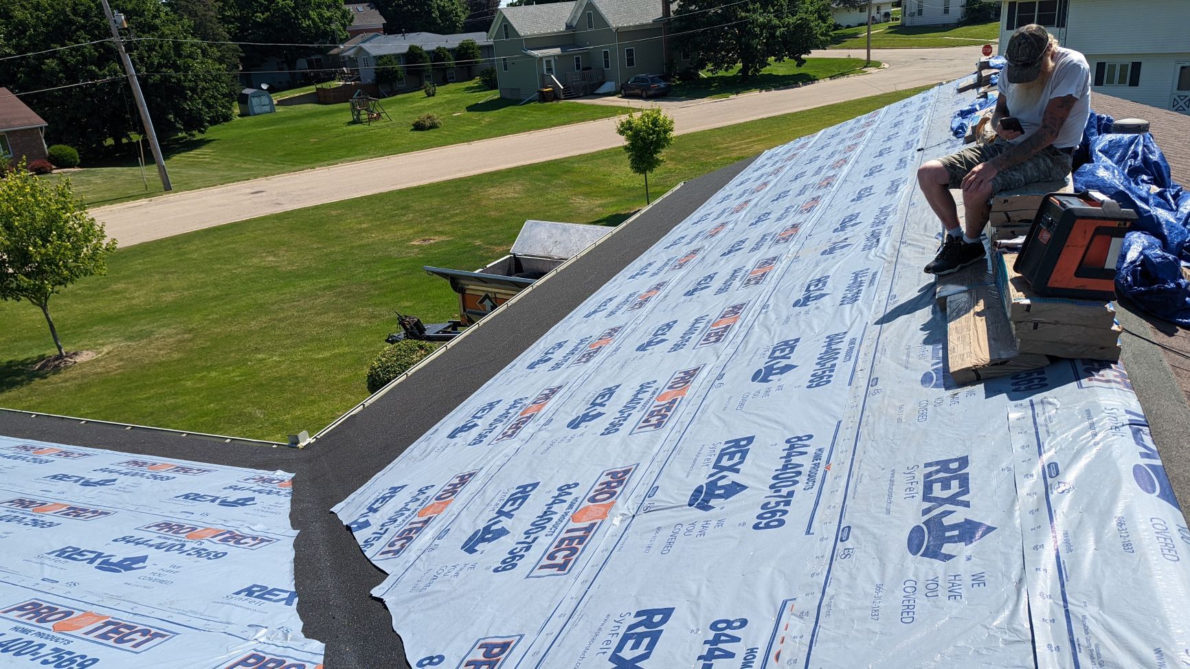 Person installing shingles on a roof covered in blue underlayment, residential neighborhood in background.
