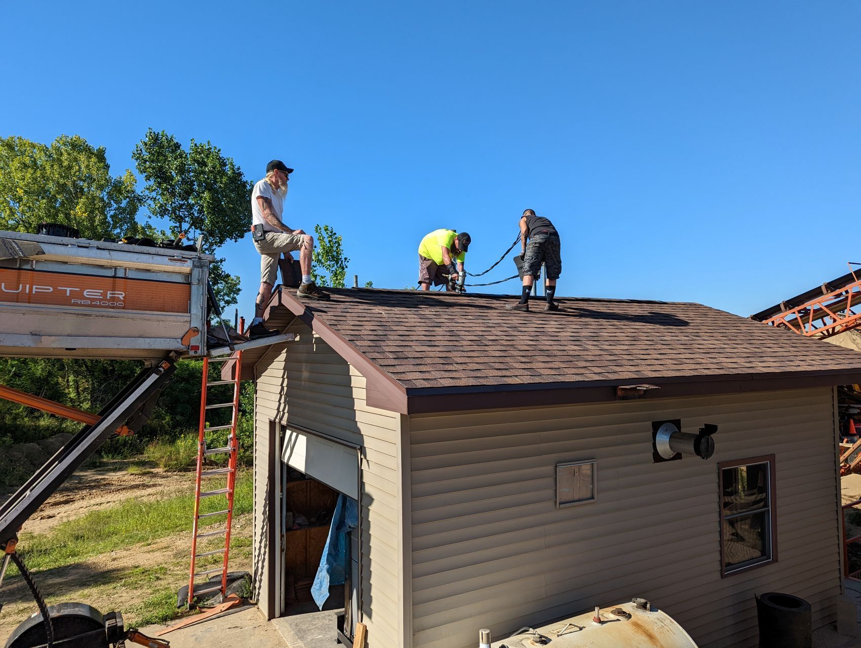 Three workers on a brown-shingled roof under a blue sky, repairing a small, beige building.